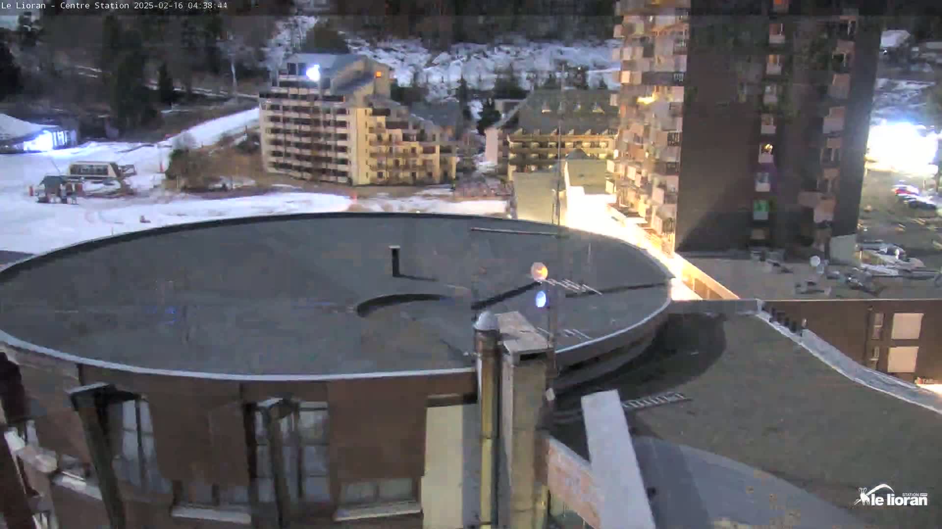 A snow-covered ski resort at night is seen from a rooftop, showing a circular building in the foreground and several multi-story buildings in the background.