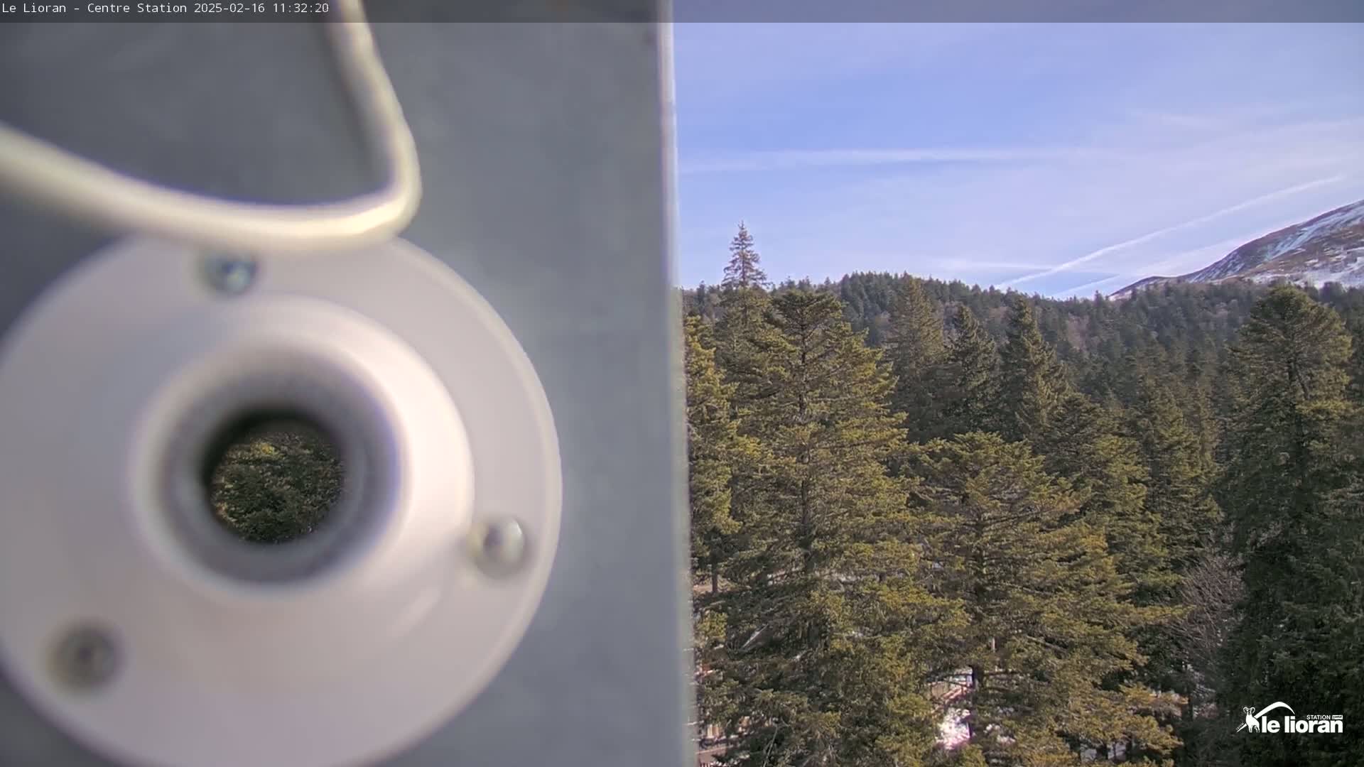 A camera lens partially obstructs a view of a coniferous forest under a mostly clear, sunny sky with a few clouds.