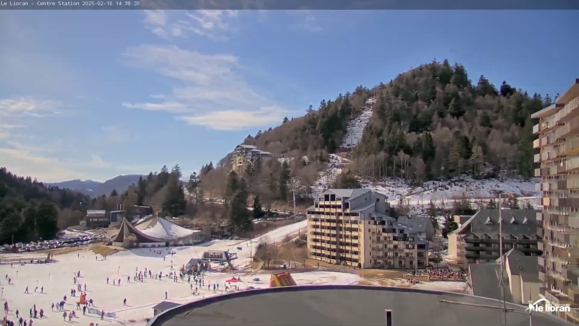A snowy ski resort with several buildings, a ski slope with people skiing and snowboarding, and a partly cloudy sky.