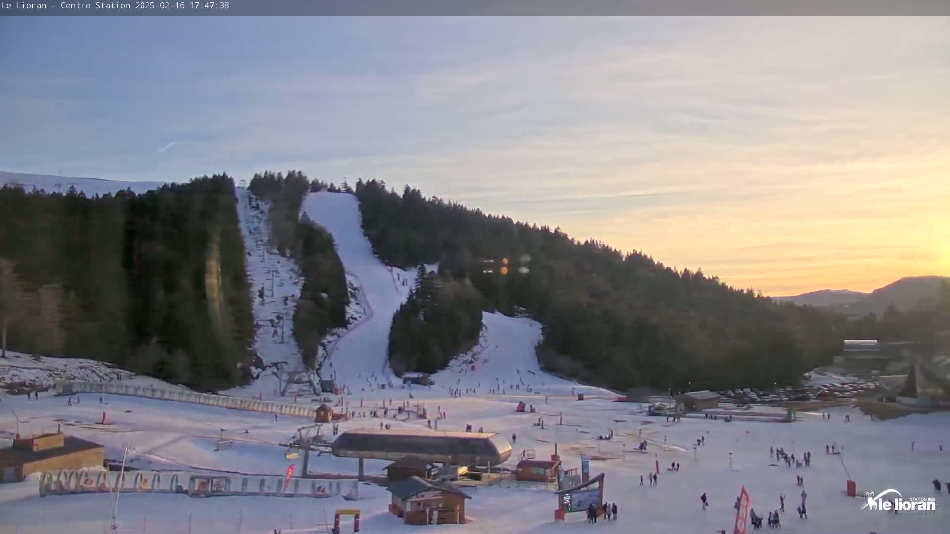 A ski resort at sunset, with snow-covered slopes and skiers visible, under a mostly clear sky.