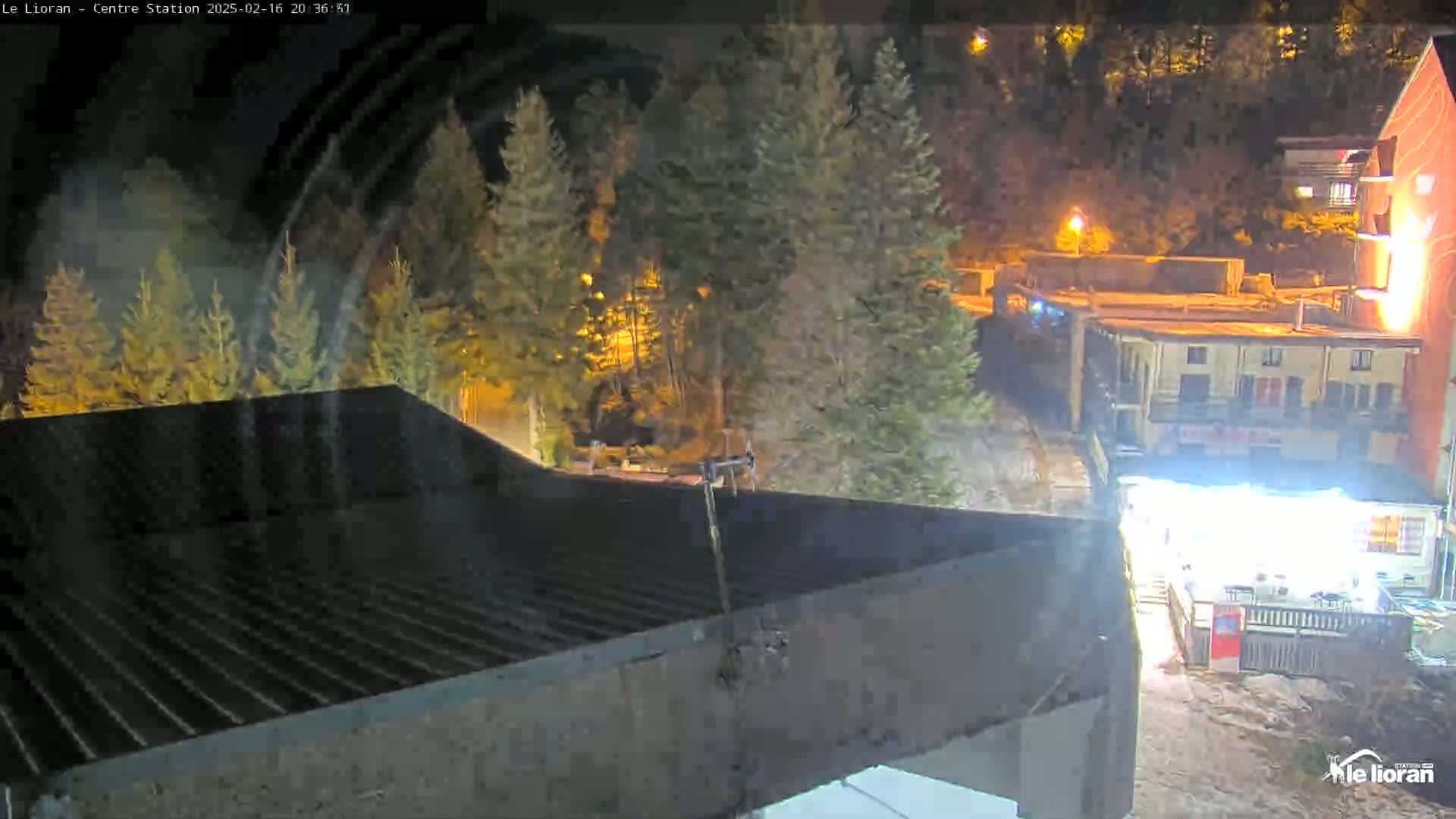A nighttime view of a dark, sloped roof overlooking illuminated buildings and evergreen trees under a clear sky.