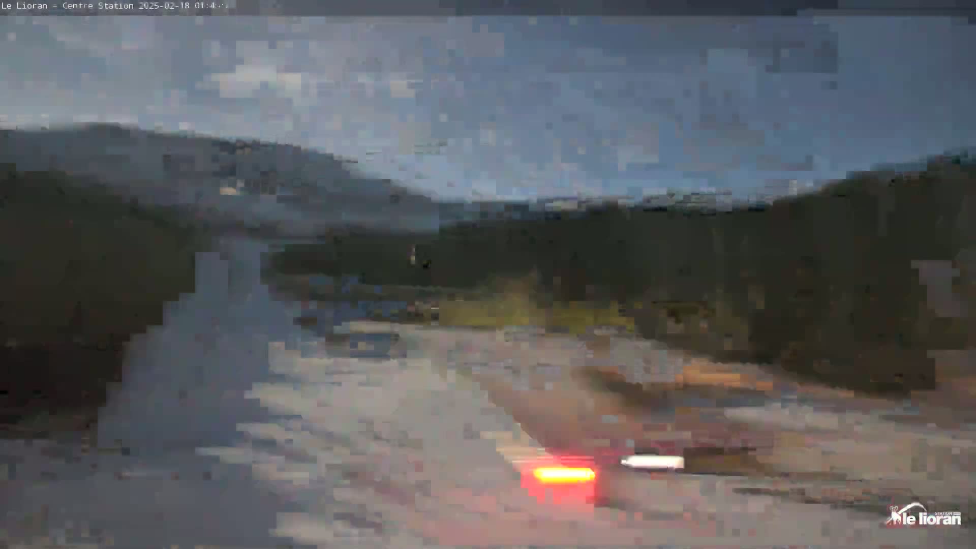 A blurry view of a road at dusk or dawn, with dark hills and a partly cloudy sky in the background, and a vehicle with its taillights on visible in the foreground.