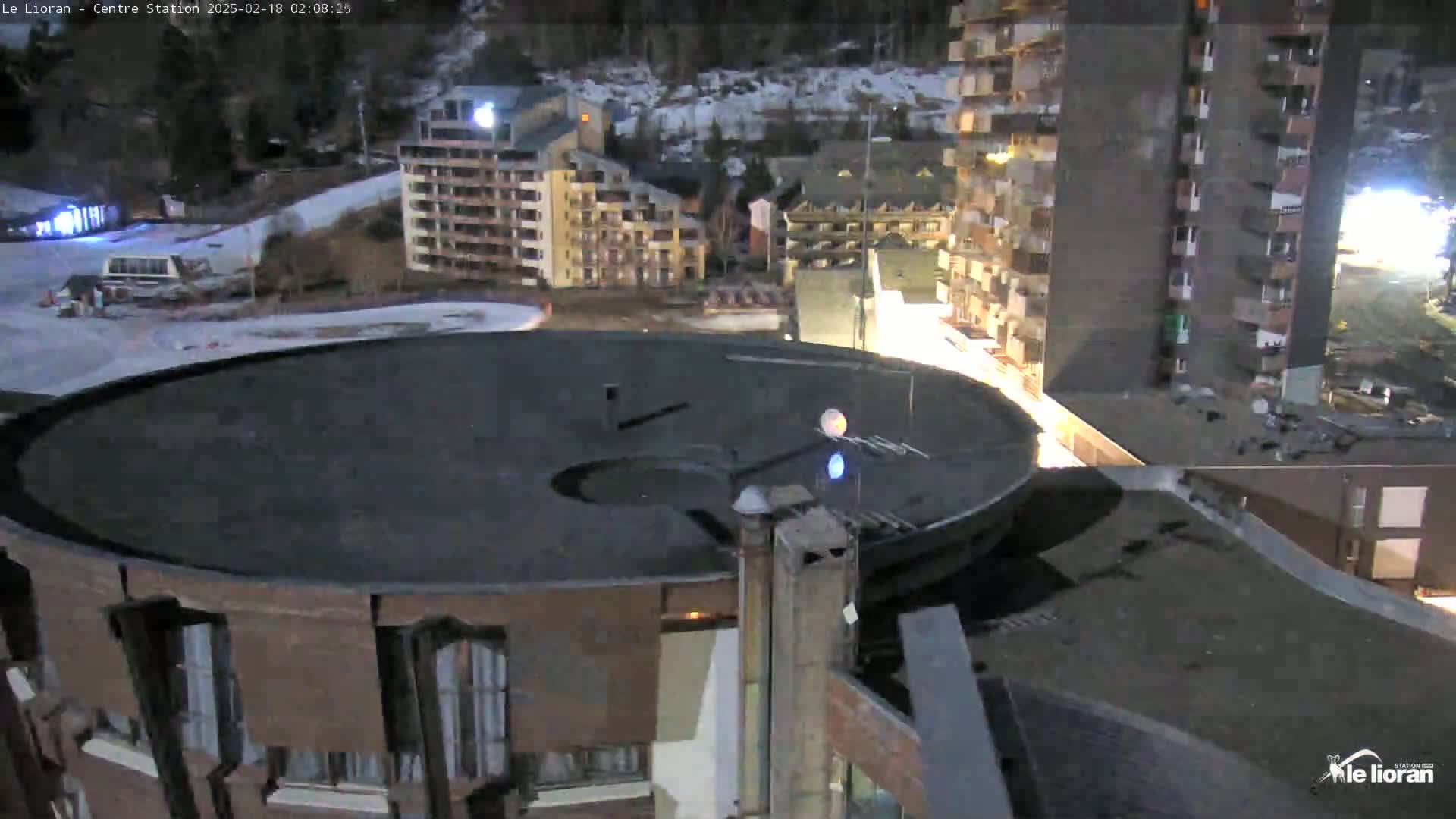A nighttime, high-angle view shows a large, dark-colored circular roof structure next to a tall apartment building, with other buildings and a snowy landscape visible in the background.