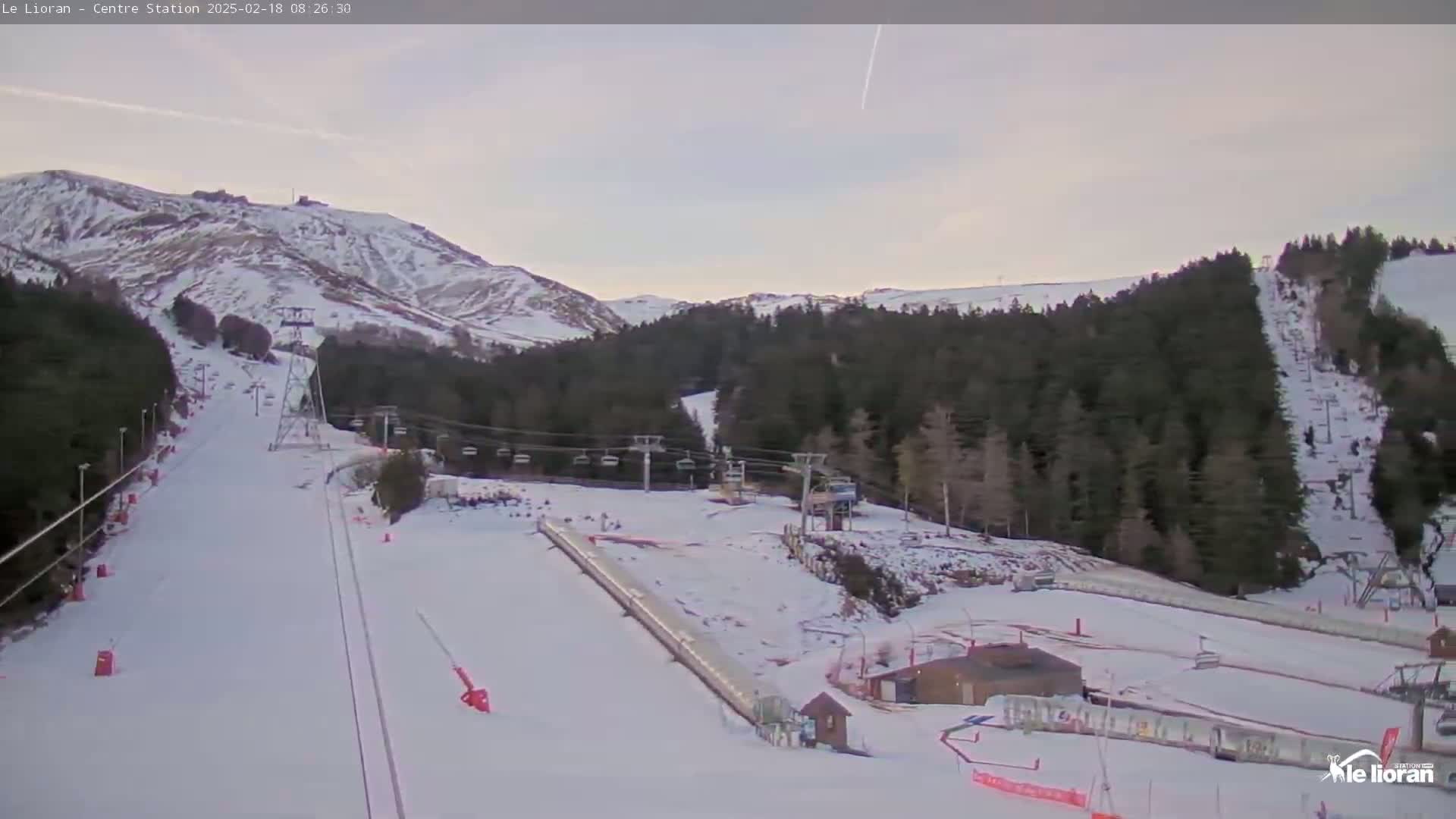 A snow-covered ski slope with chairlifts and a mountain in the background under a partly cloudy sky.