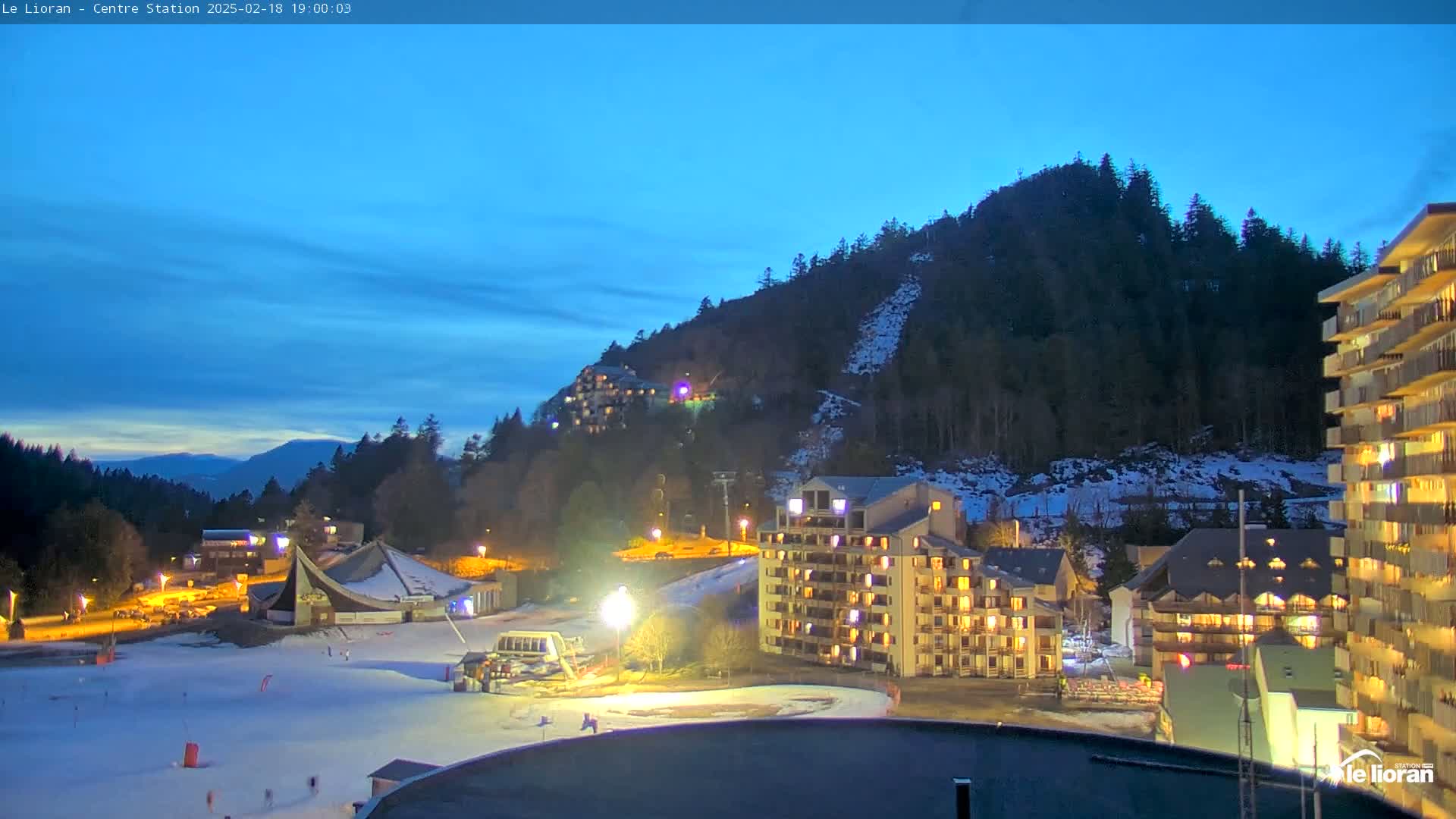 A nighttime view of a snow-covered ski resort with illuminated buildings nestled against a dark, snow-dusted mountainside under a twilight sky.