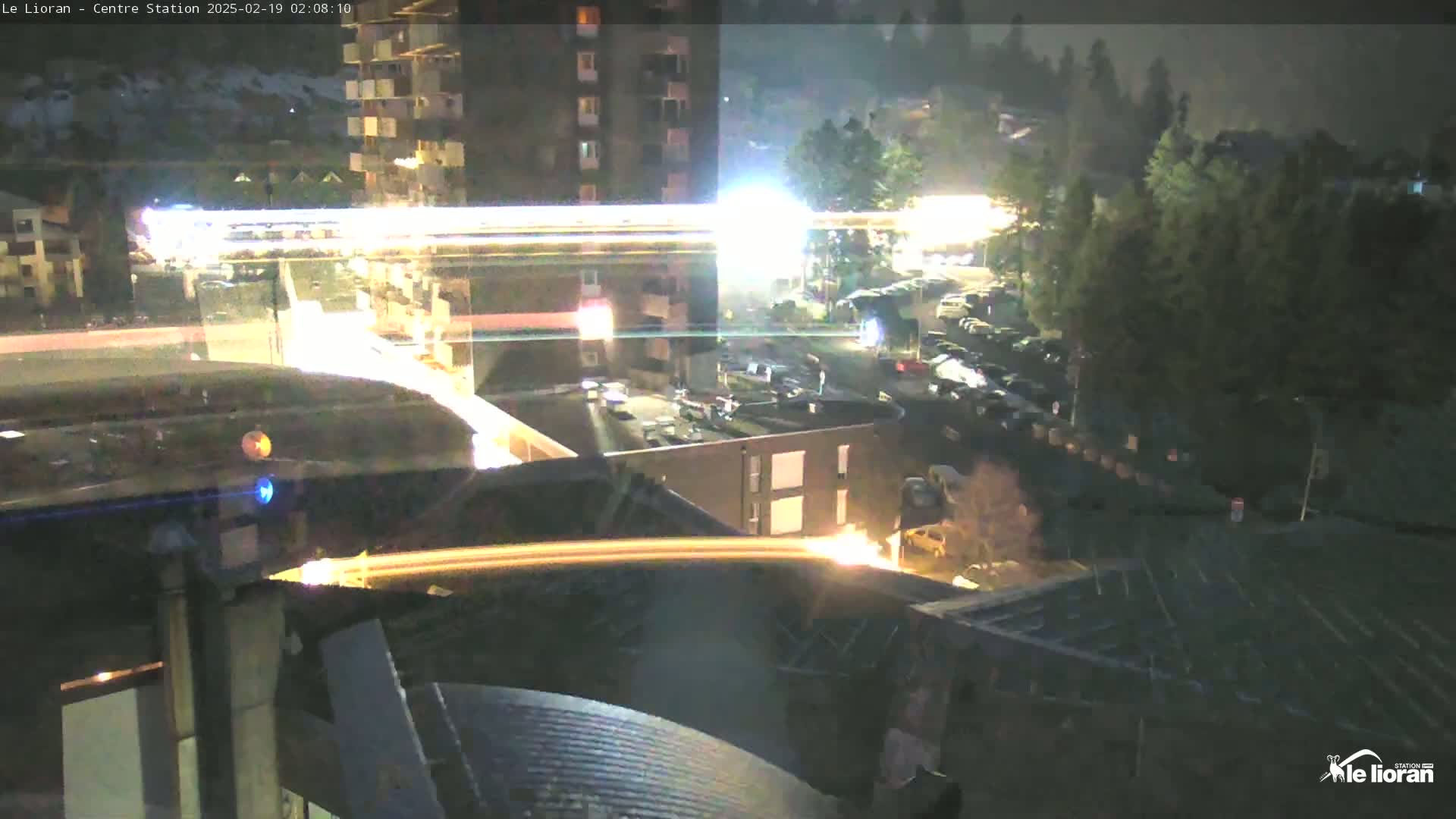 A nighttime aerial view of a snow-covered mountainside, buildings, and a parking lot with light streaks from moving vehicles.