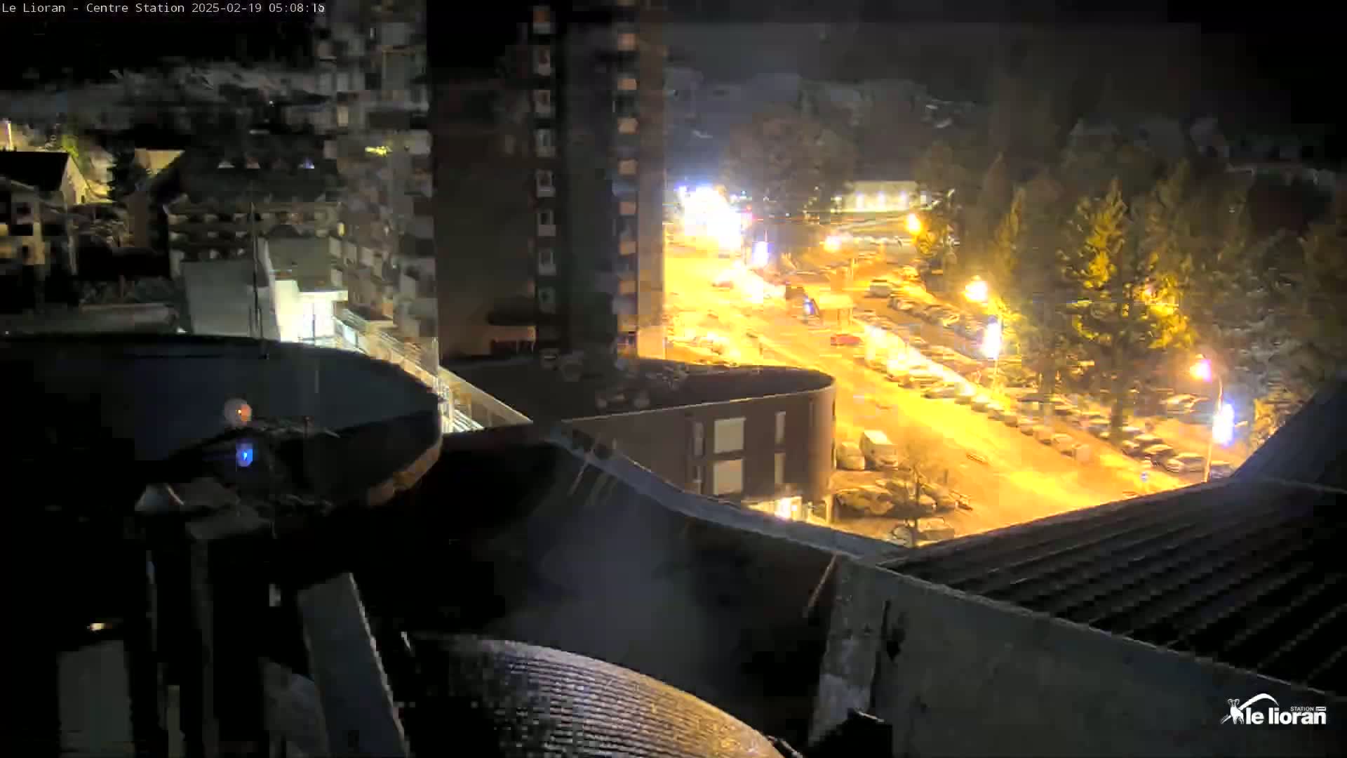 A nighttime, high-angle view of a snow-covered street with parked cars and illuminated buildings, seen from a rooftop.