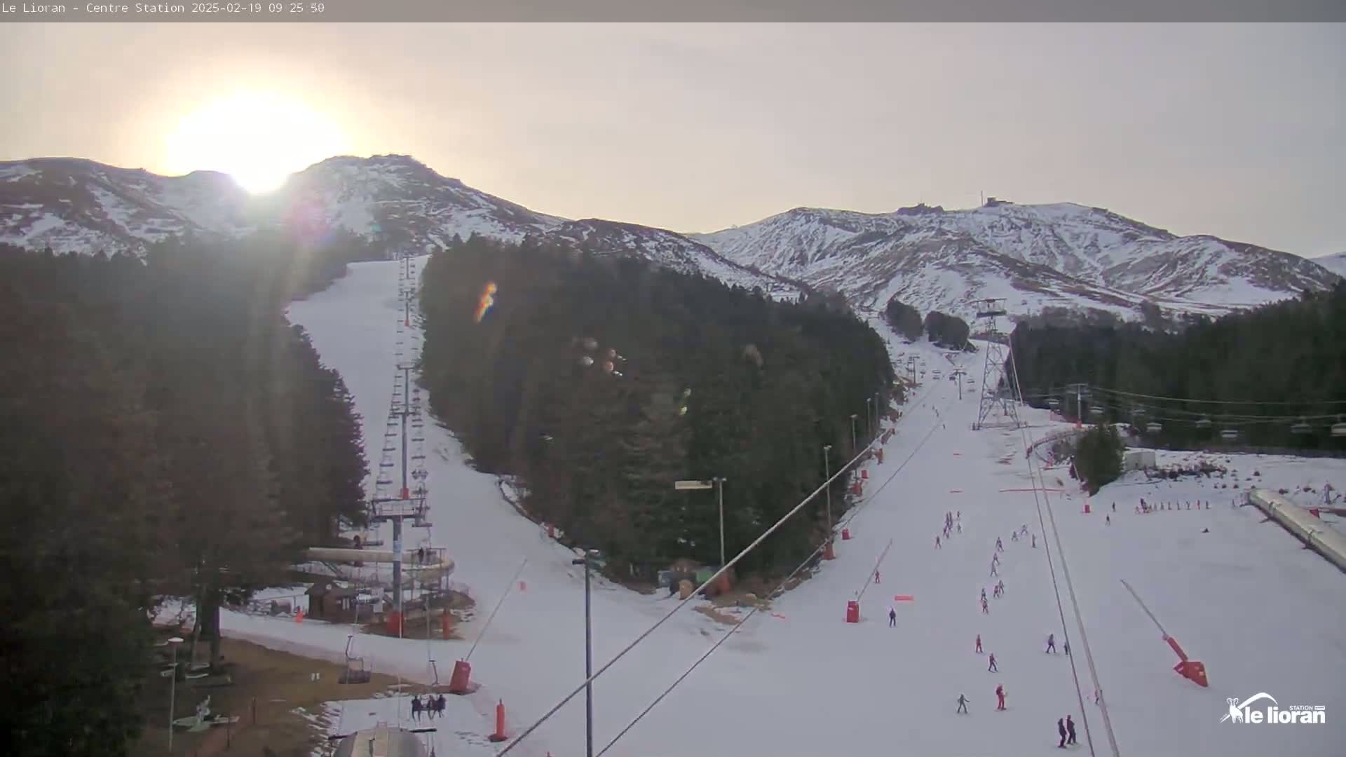 A sunlit snowy ski slope with several skiers and a chairlift, surrounded by snow-dusted mountains and evergreen trees under a mostly clear sky.