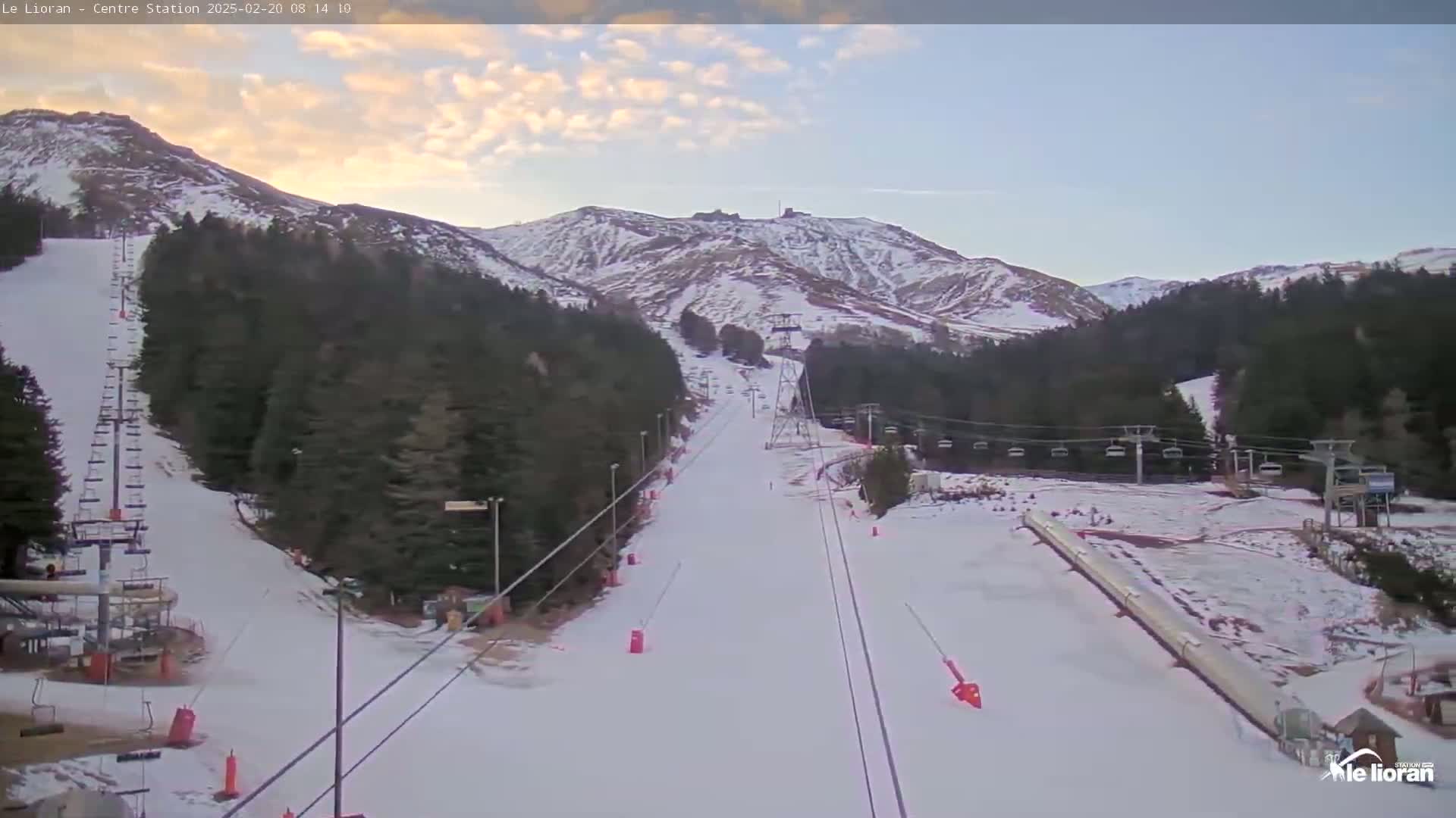 A snow-covered ski slope winds down a mountainside under a partly cloudy sky, with ski lifts and sparsely populated runs visible.