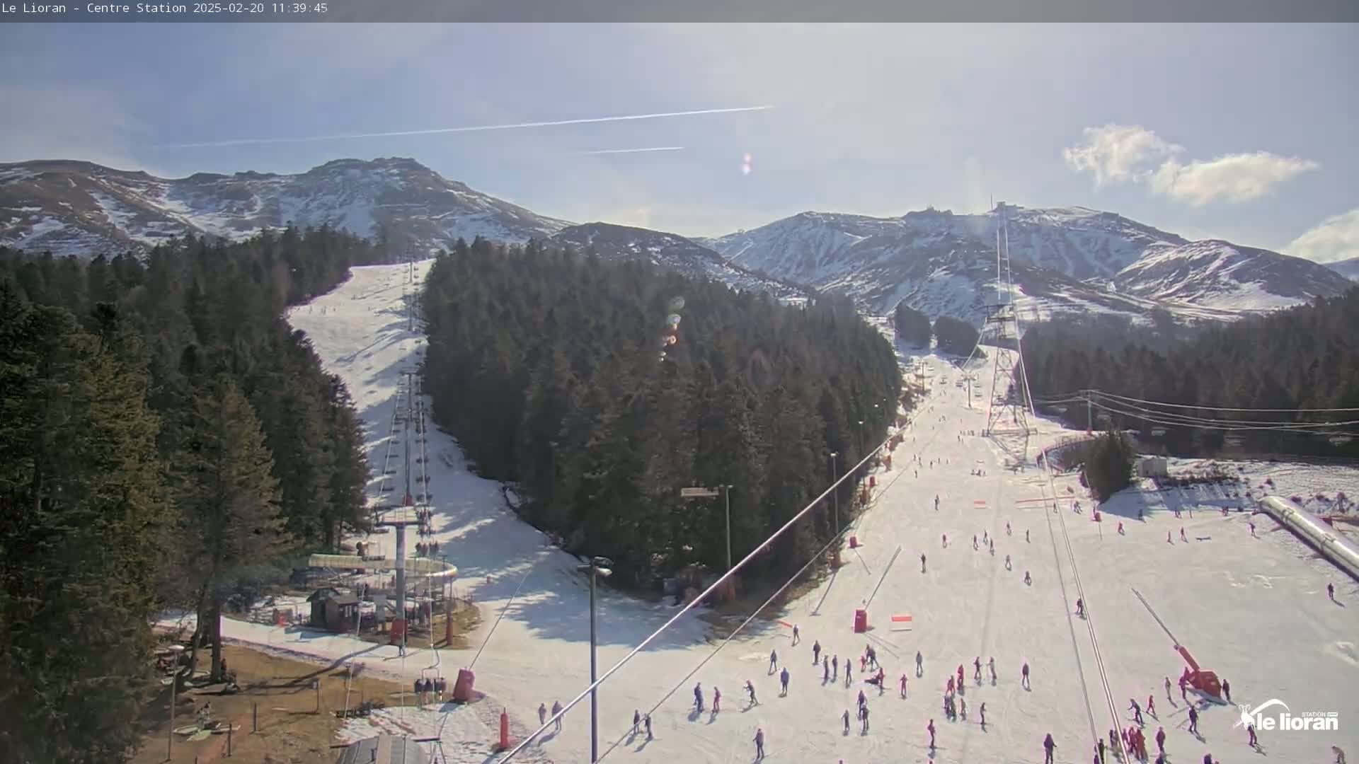 A sunny, snow-covered ski resort with skiers and snowboarders on the slopes, surrounded by evergreen forests and snow-capped mountains.