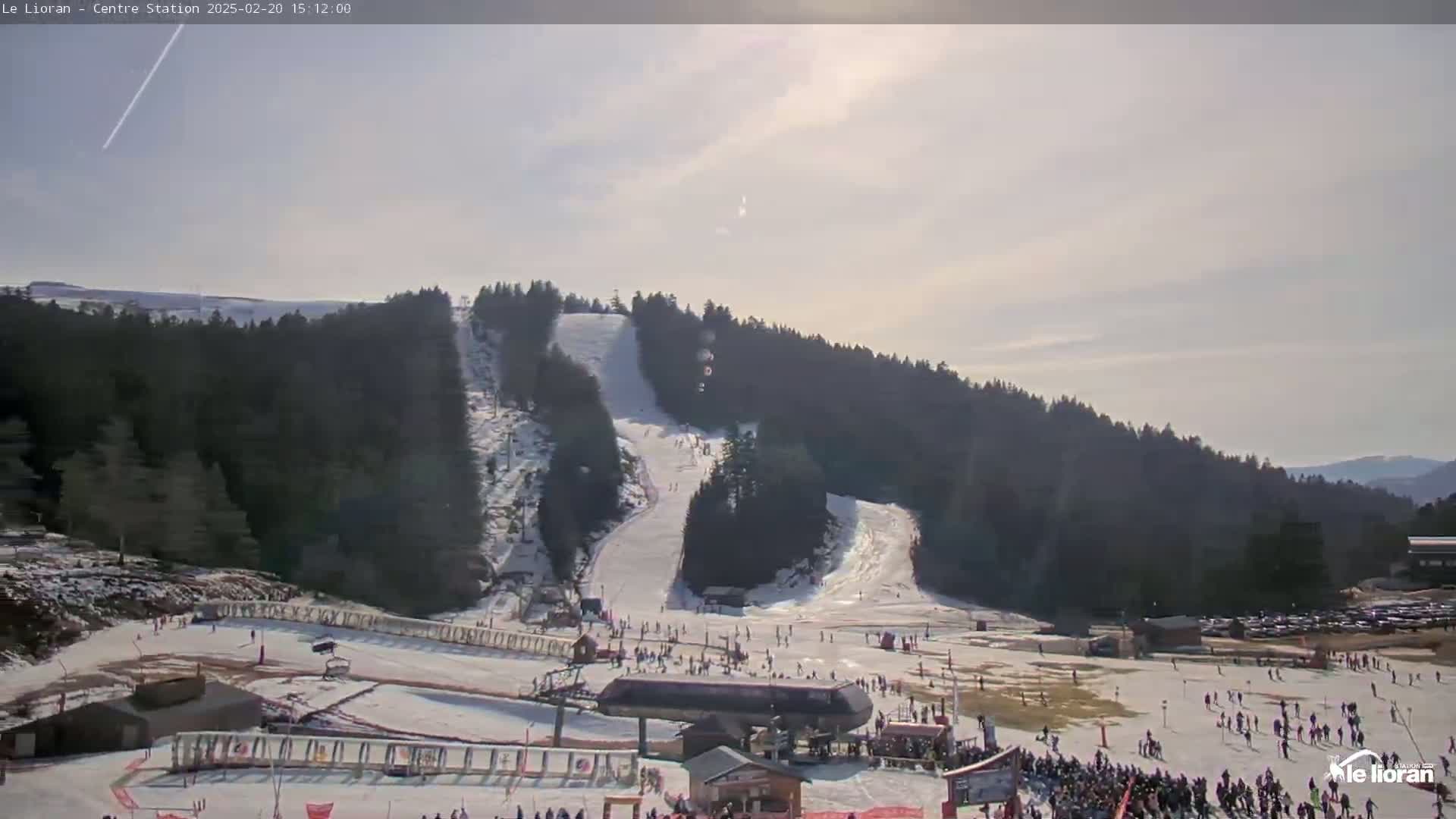A sunny day at a snow-covered ski resort shows several ski slopes with skiers and snowboarders, a ski lift, and a parking lot.