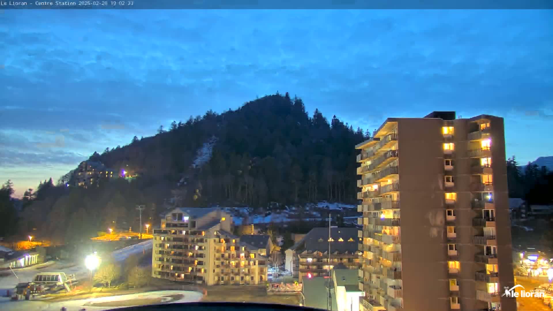 A twilight view of a mountain town shows several lit-up apartment buildings at the base of a snow-dusted mountain under a clear, blue-tinged sky.