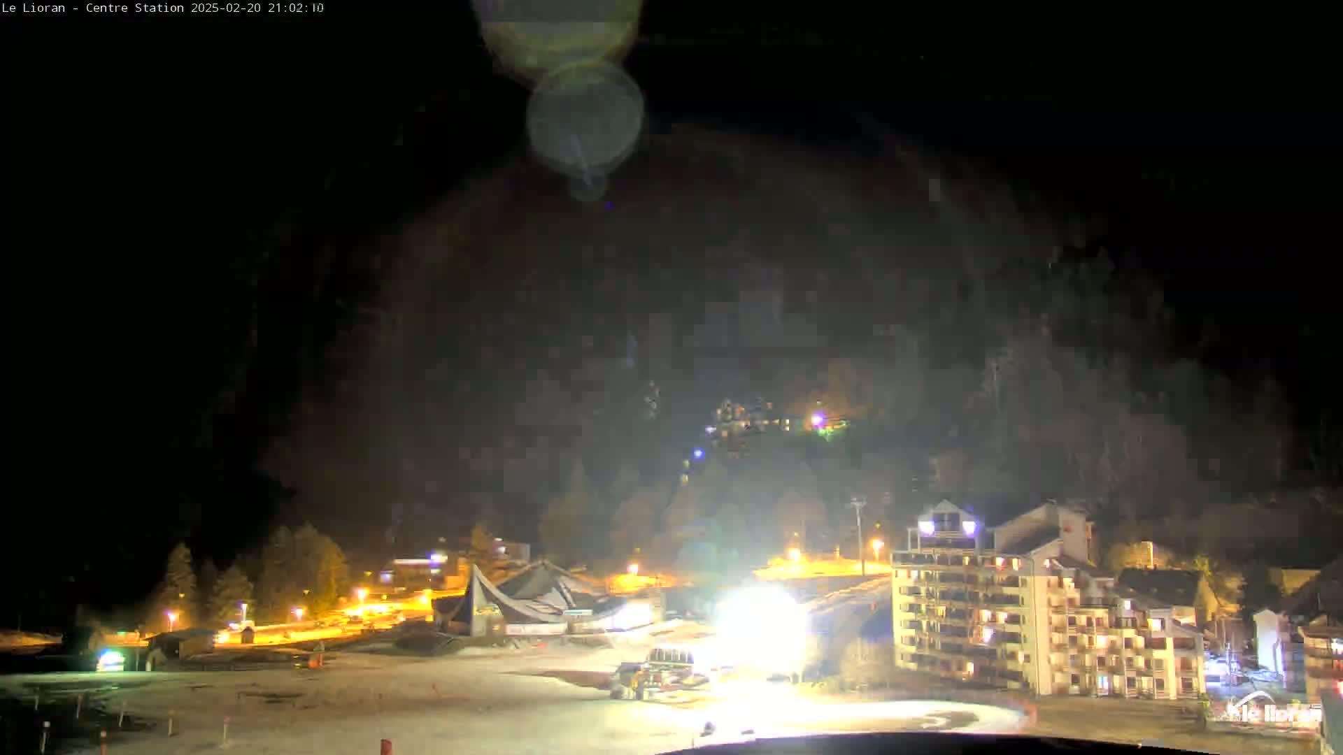 A nighttime view of a snow-covered area with buildings and a snow groomer illuminated, under a dark, cloudy sky.