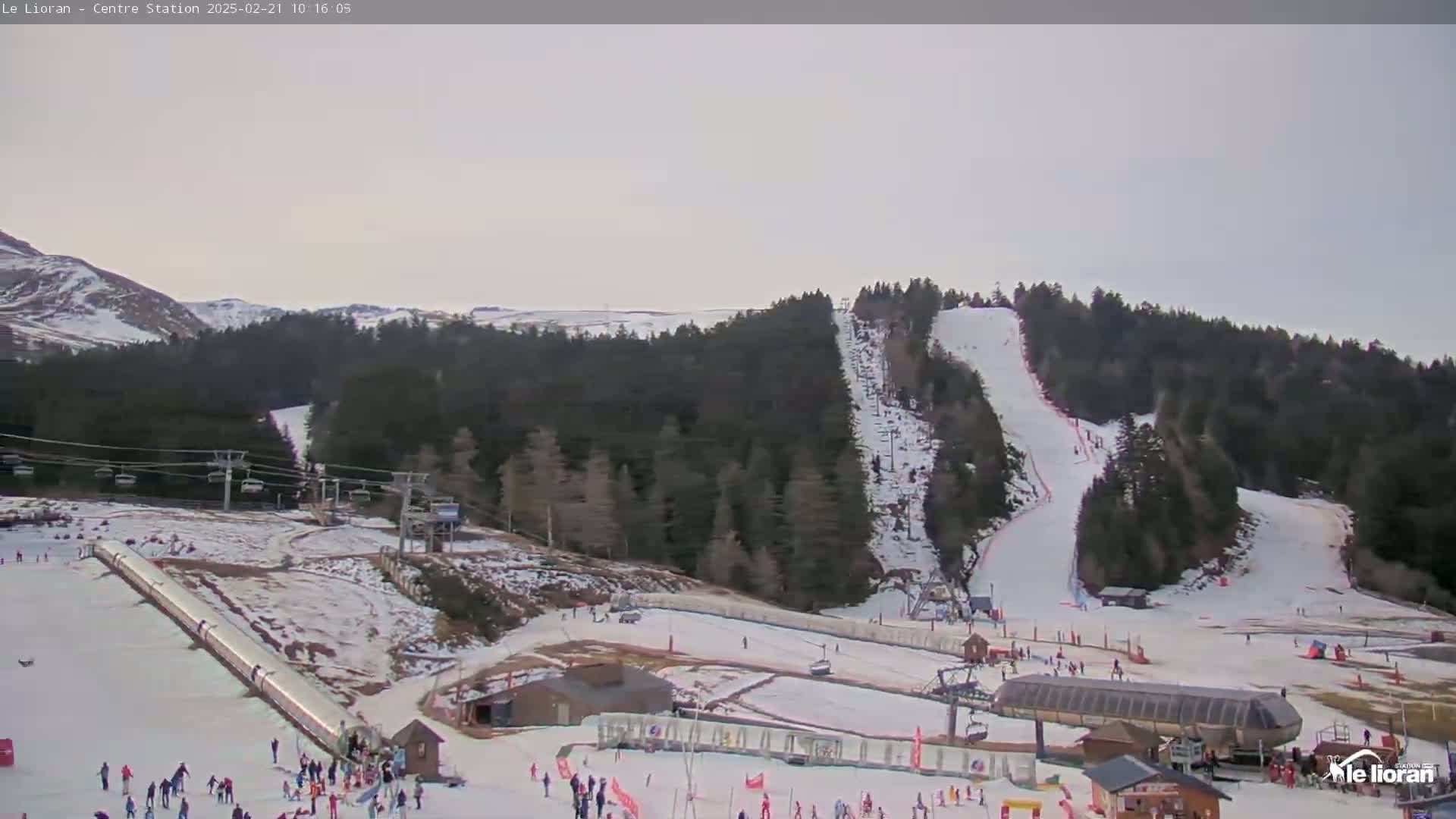 A snowy ski resort with several ski slopes, chairlifts, and skiers visible on a mostly cloudy day.