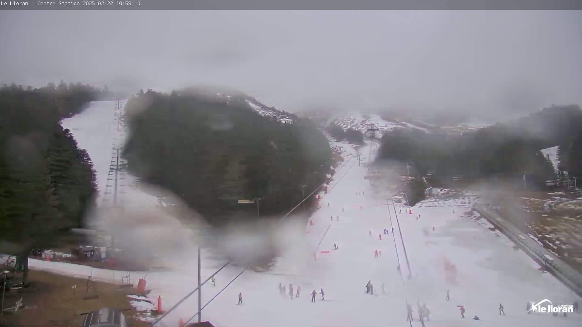 A snowy ski slope with several skiers visible, surrounded by fog-covered trees under overcast conditions.