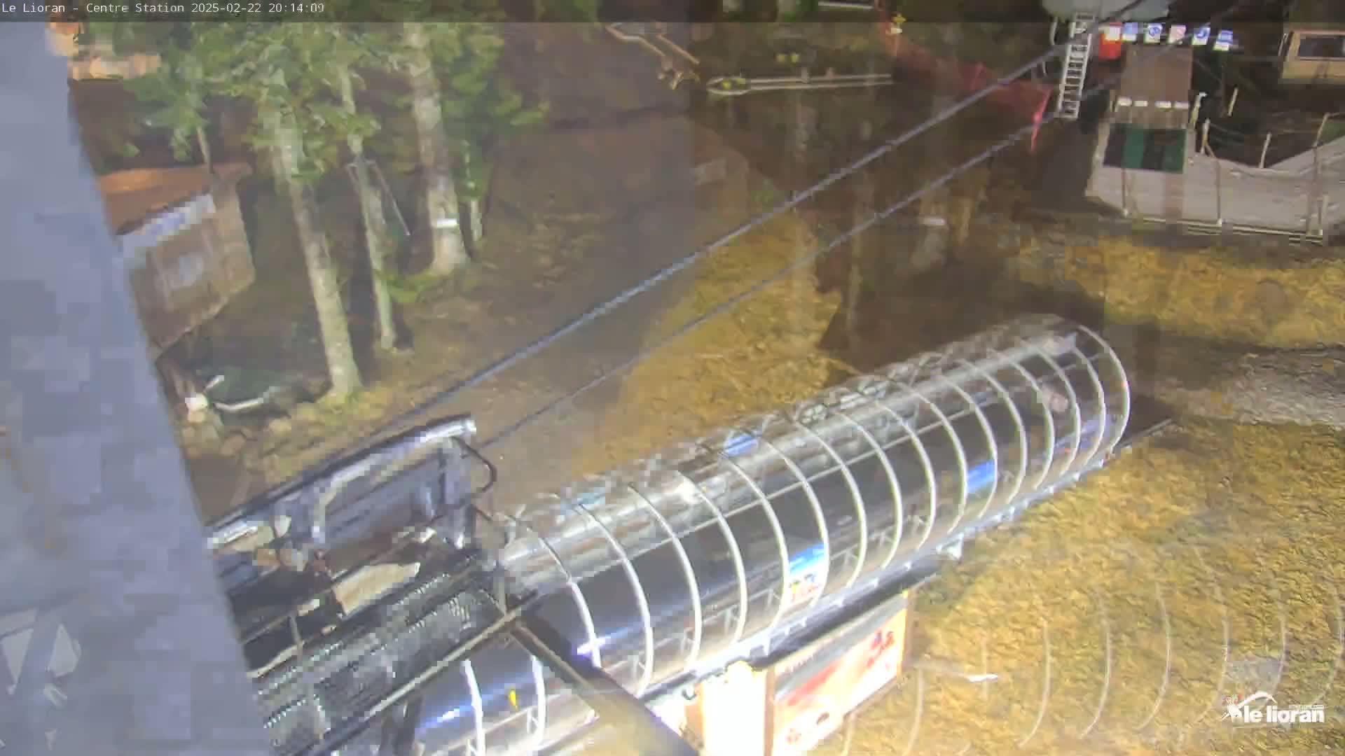 A transparent enclosed chairlift gondola moves along a cable above a wet, leaf-covered ground, surrounded by trees at night.