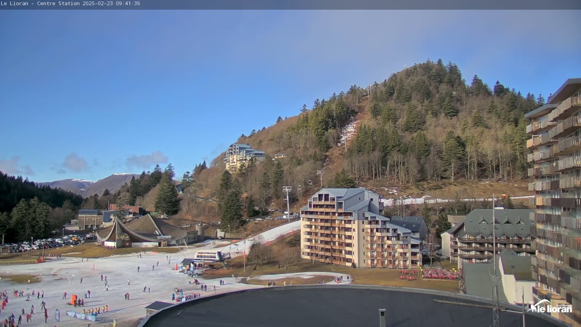 A sunny day at a ski resort shows a snow-covered slope with people skiing and snowboarding, several buildings, and a hill covered in evergreen trees.