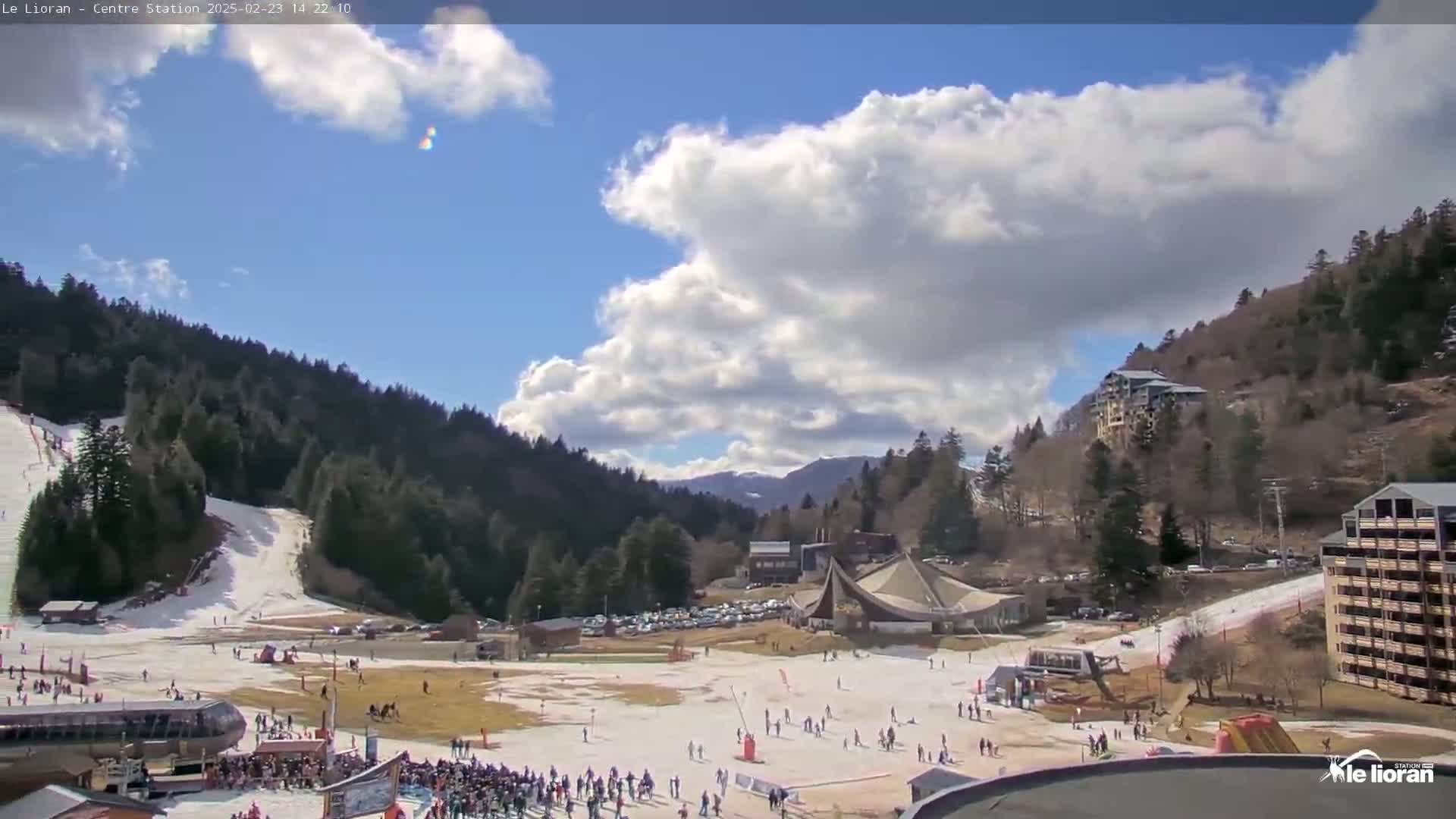 A partly cloudy sky looks down on a snow-covered ski resort nestled in a valley between forested mountains.