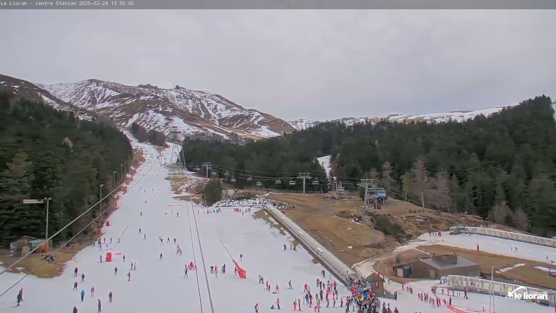A ski slope with several skiers and snowboarders on a cloudy day, surrounded by snow-dusted mountains and evergreen forests.