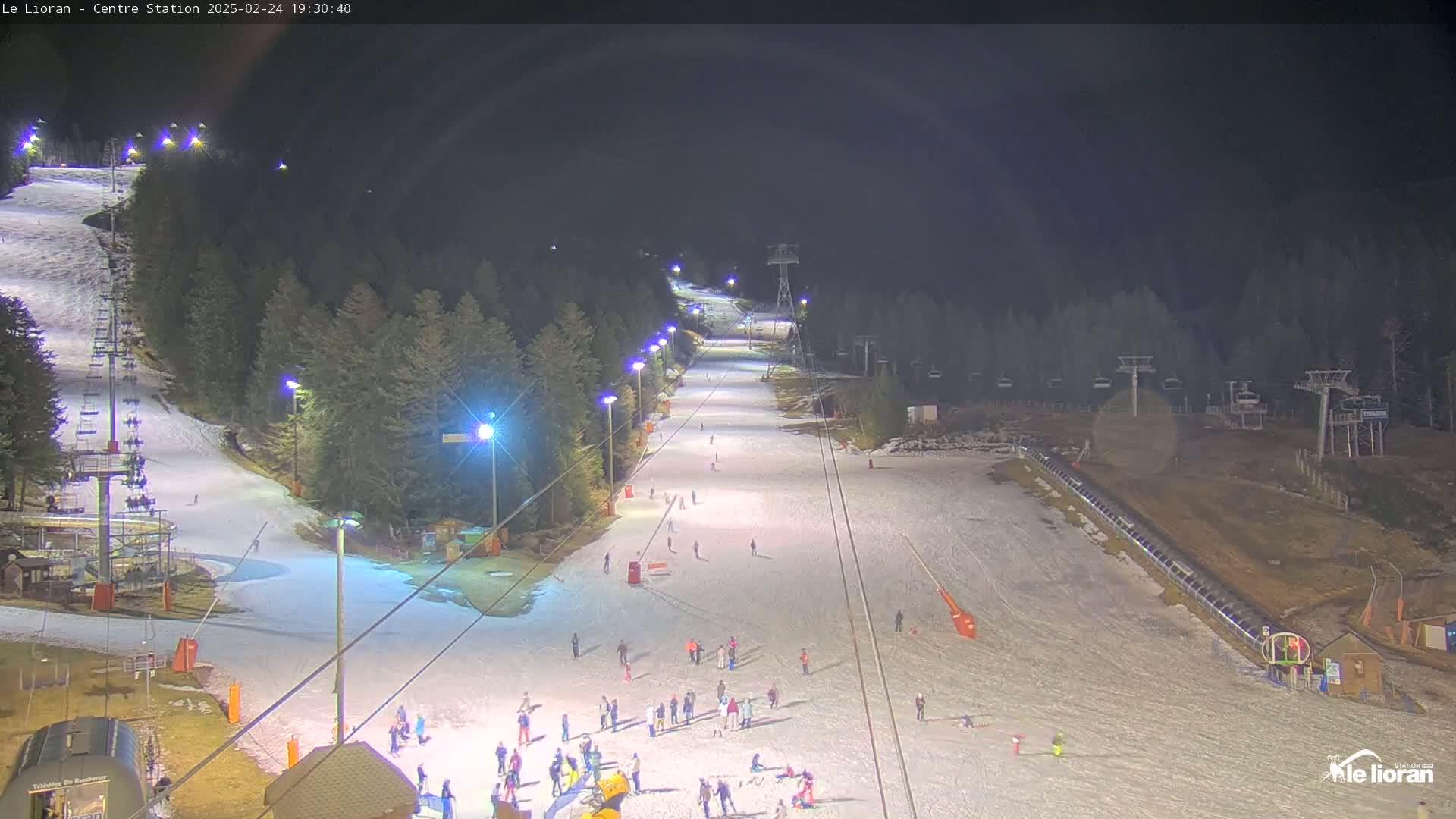 A snow-covered ski slope at night is illuminated by artificial lights, with several skiers and snowboarders visible, and ski lifts in the background.