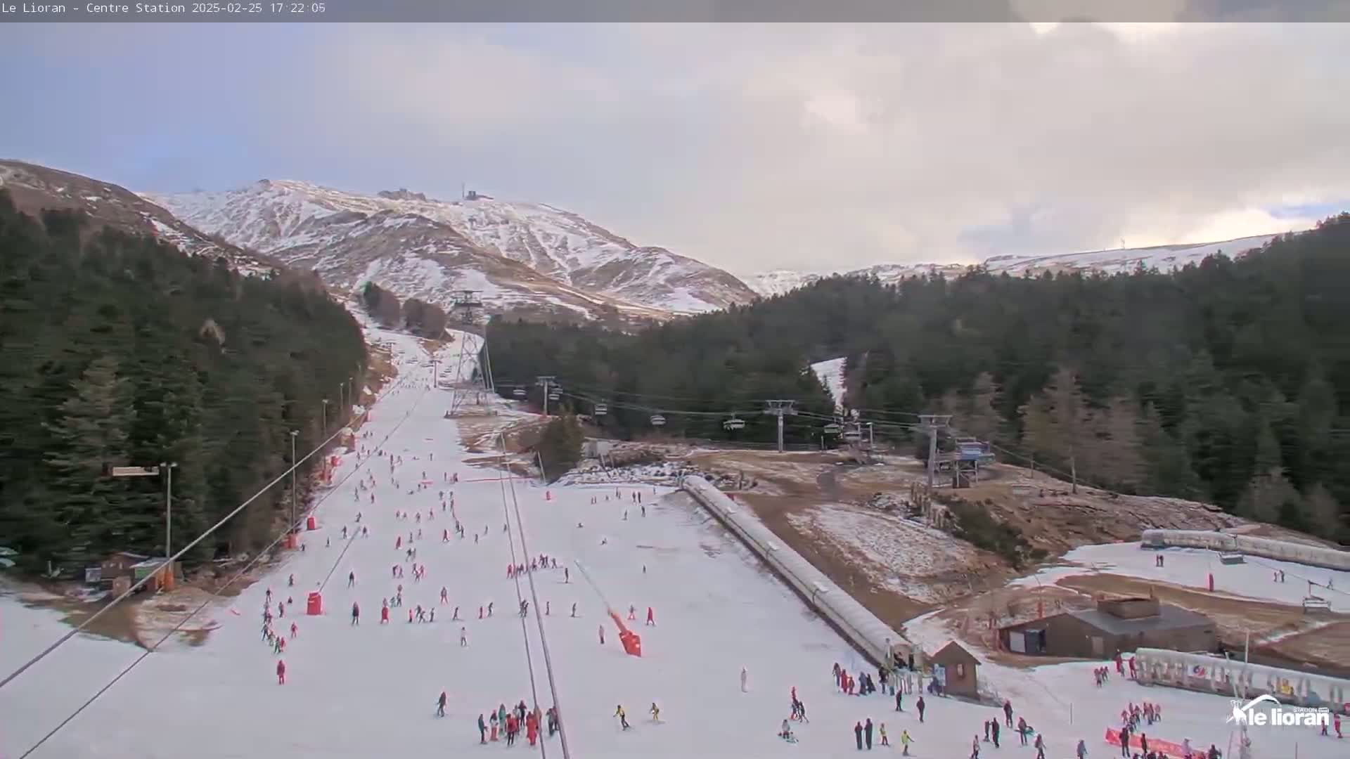 A snowy ski slope with skiers and chairlifts is nestled in a mountain valley under an overcast sky.