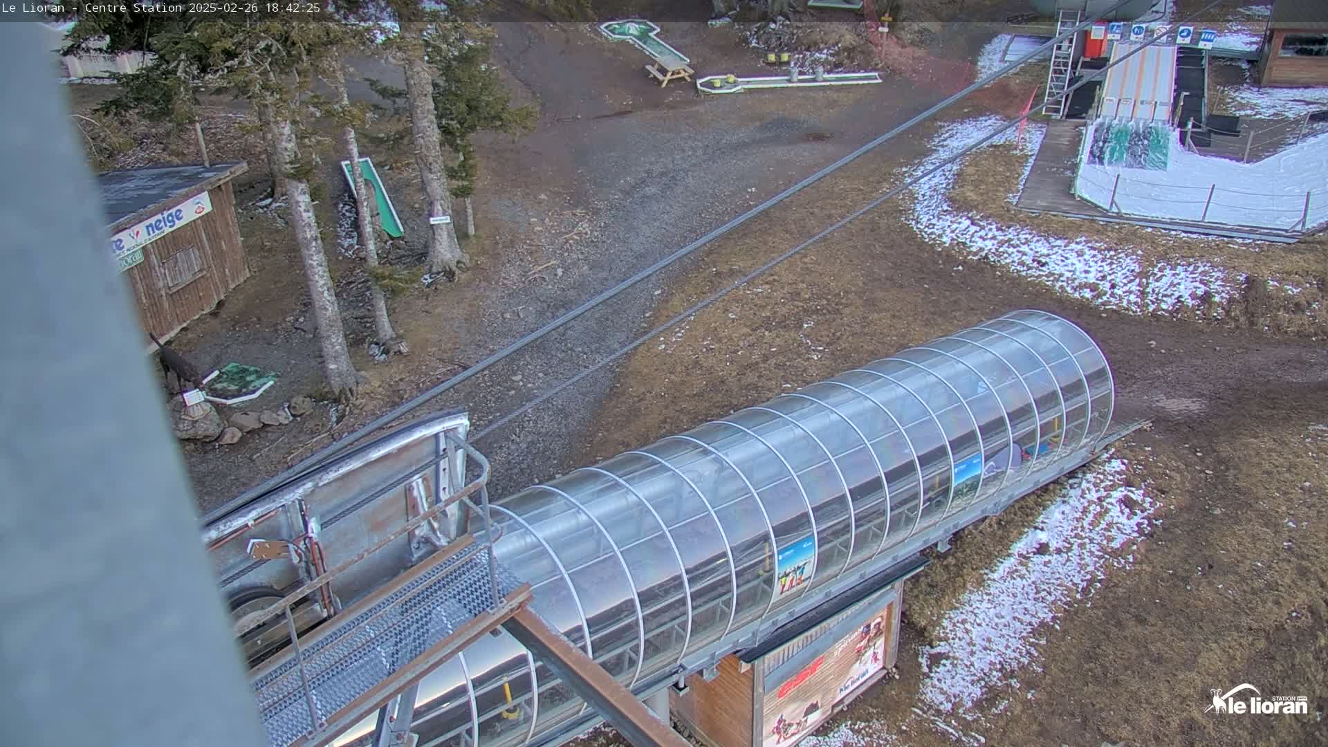 A covered ski lift is seen from above, surrounded by a mostly barren landscape with patches of snow, some trees, and mini golf courses.