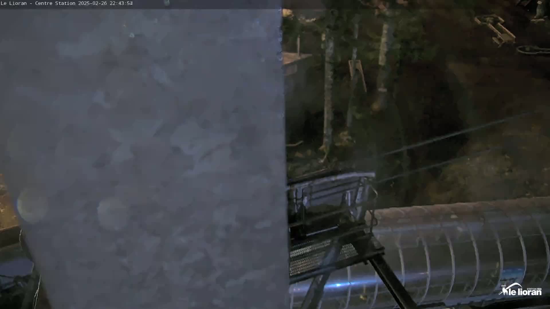 A nighttime view shows a dark-colored structure partially obscuring a chairlift next to a transparent structure, with trees in the background under dark conditions.
