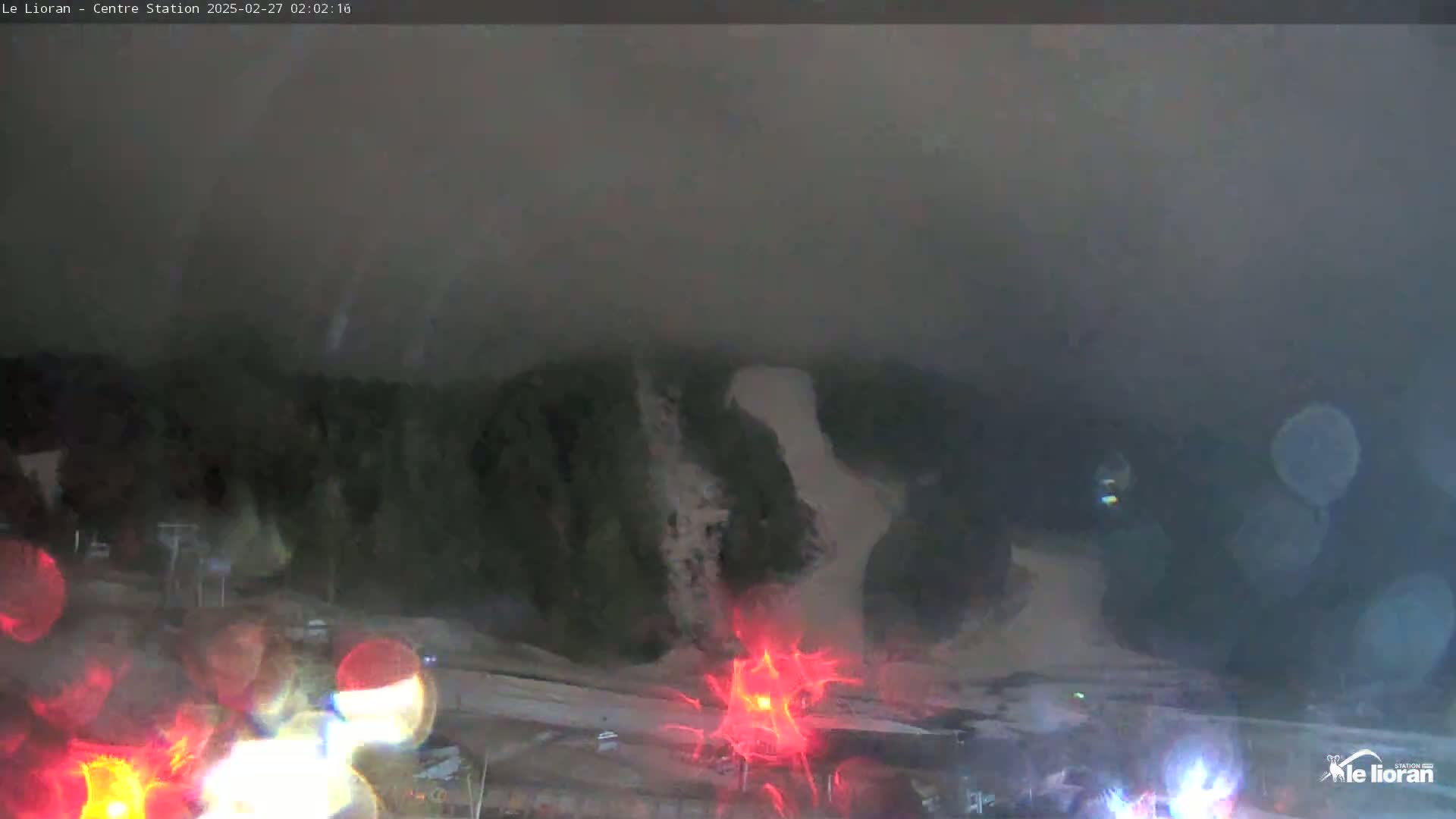 A nighttime view of a snow-covered ski slope, seen through rain-covered glass, with faint lights visible in the distance.