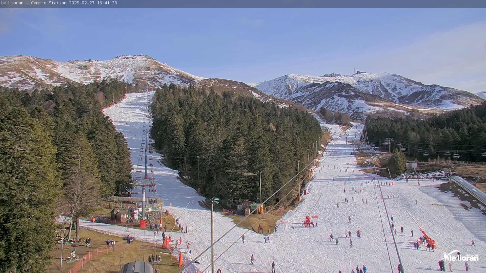 A ski slope with several skiers and snowboarders on it, nestled in a snow-dusted mountain valley under a clear blue sky.