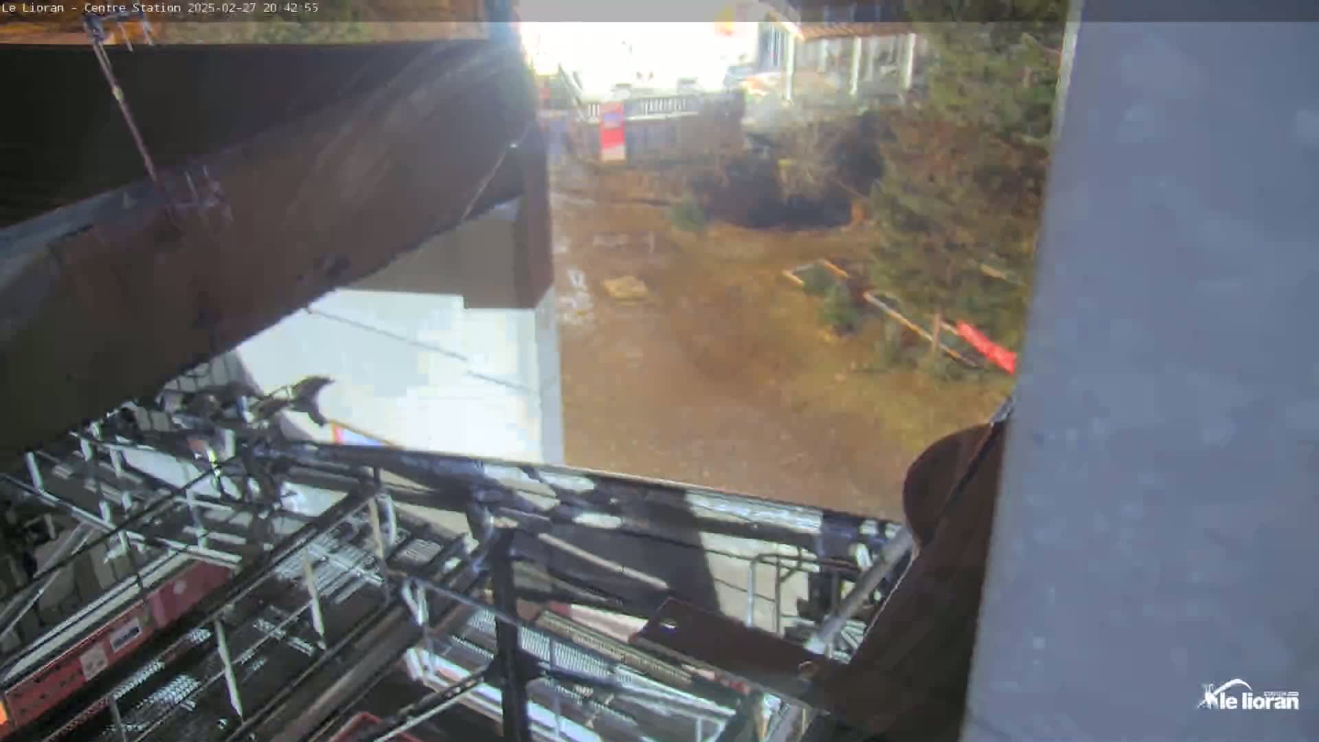 A view from inside a gondola shows a wet, muddy ground with some vegetation and buildings in the background.