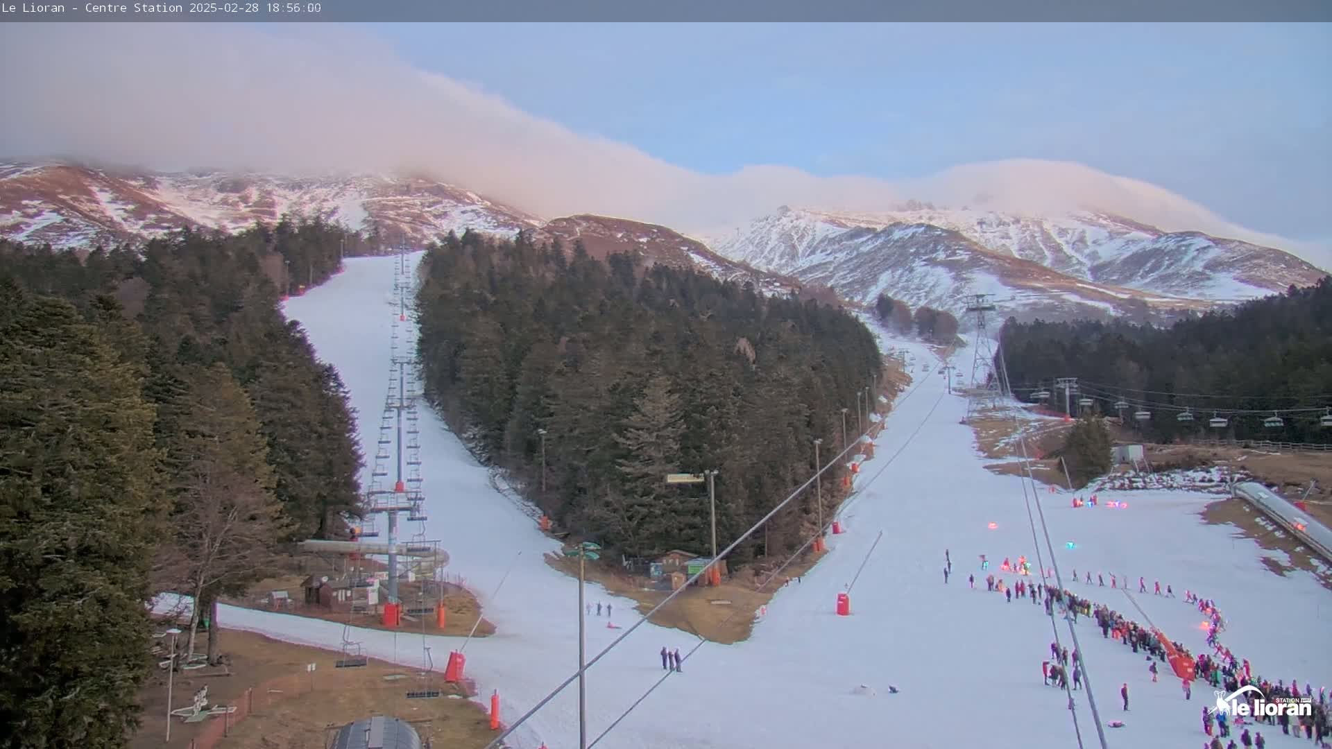 A snow-covered ski slope with several chairlifts winds down a mountainside under a partly cloudy sky.