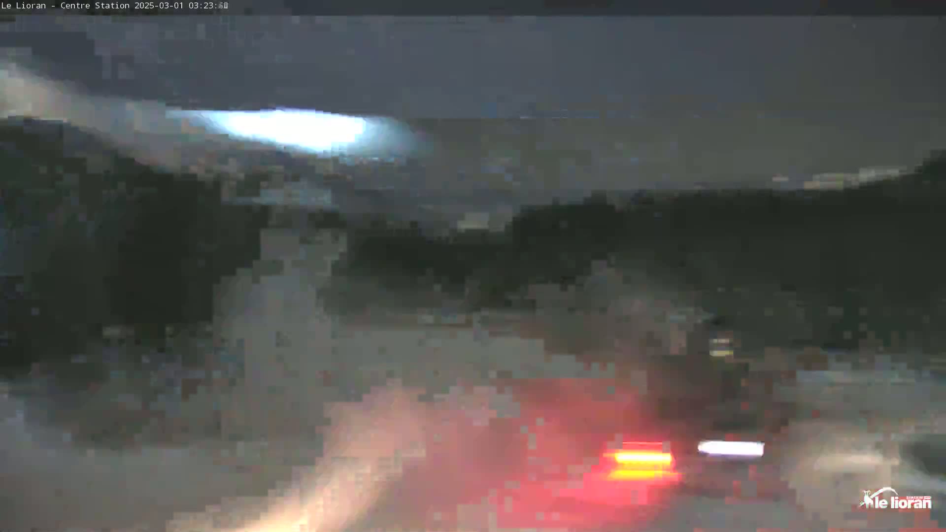 A nighttime, snowy scene shows the blurry red taillights of a vehicle in the foreground, with a dark, snow-covered landscape in the background under a dimly lit sky.
