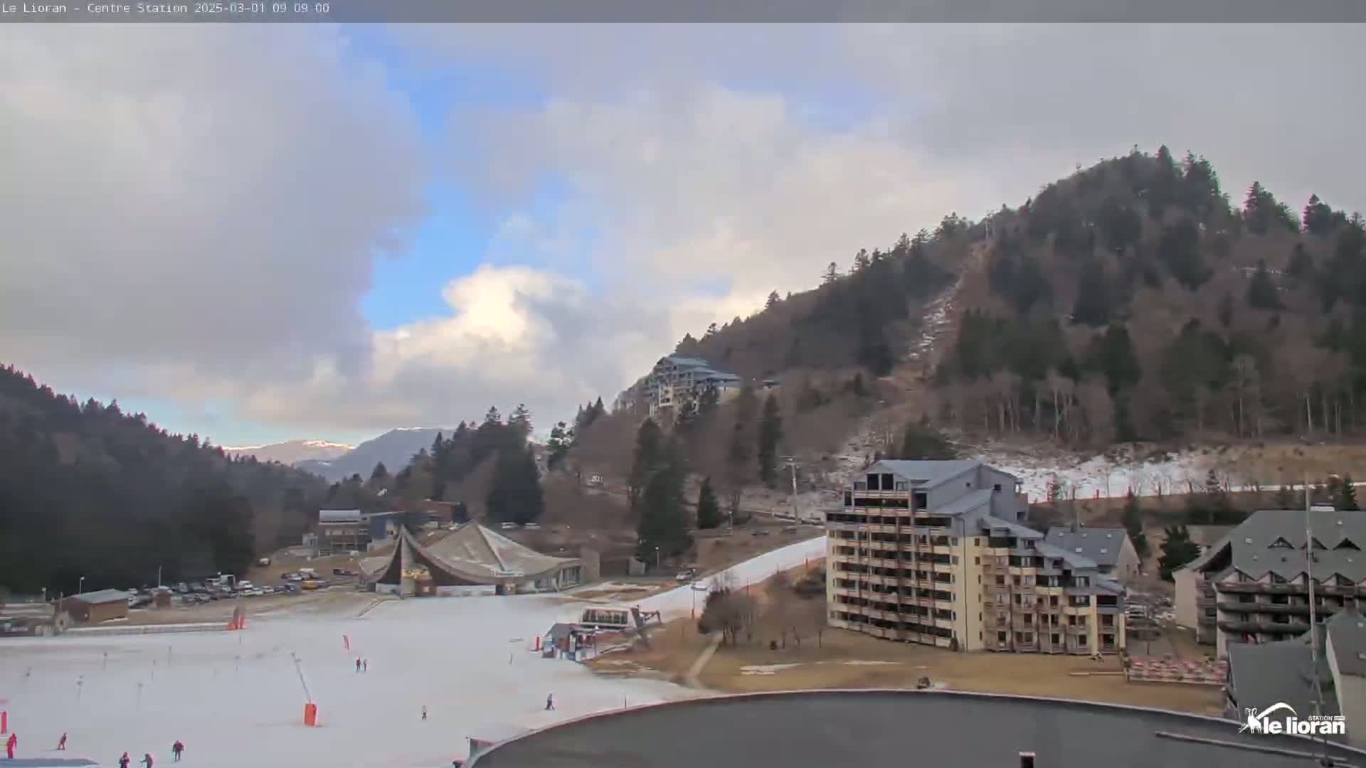 A partly cloudy day reveals a snowy ski resort nestled in a mountainside, with several buildings and a ski slope visible.