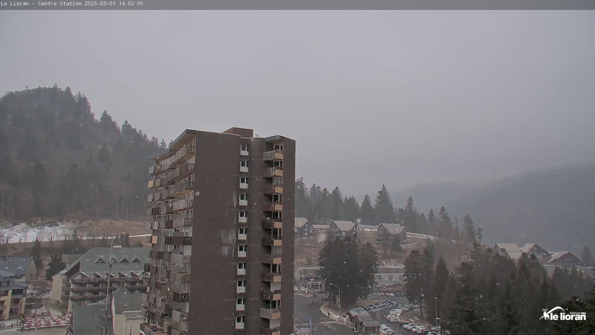 A multi-story building is seen in the foreground with light snowfall over a mountainous area covered in coniferous trees and other buildings.