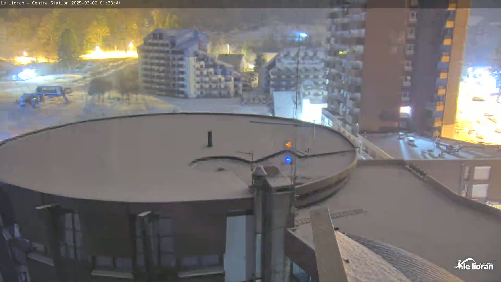 A snow-covered circular rooftop is visible, overlooking a snowy landscape with buildings and faint lights under a nighttime snowfall.