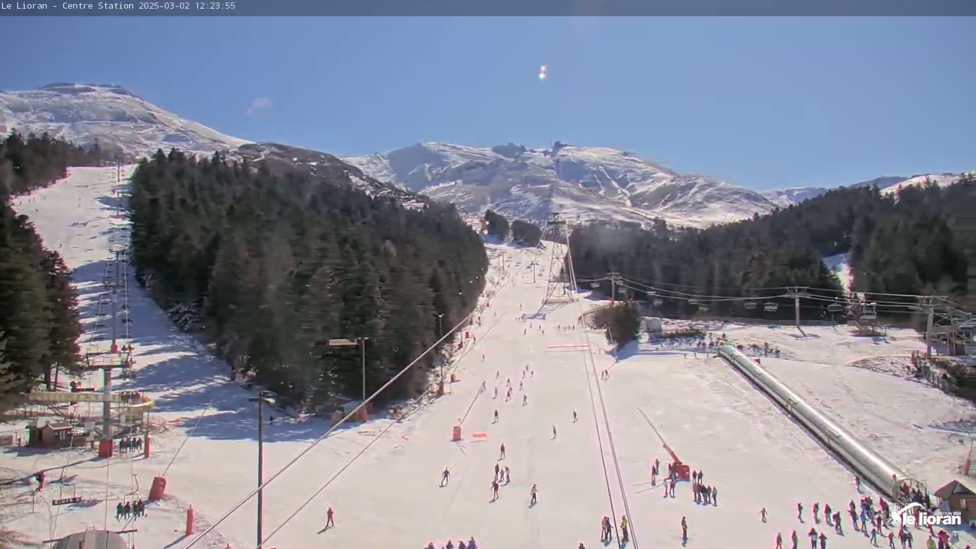 A sunny day at a snow-covered ski resort shows several skiers and snowboarders on a slope with ski lifts and a mountain in the background.