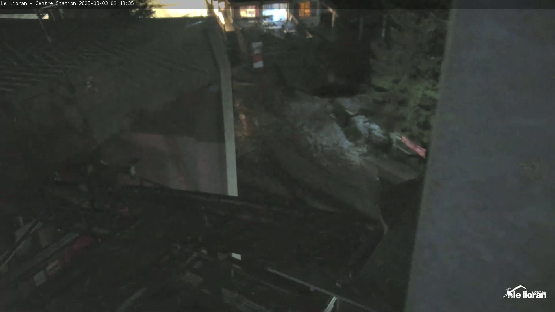 A nighttime, low-light view shows a dark, possibly snowy or icy, ground with some vegetation and structures in the background.