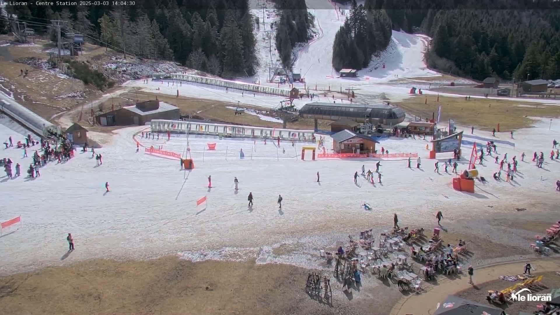 A ski resort on a sunny day with skiers and snowboarders on the slopes and people relaxing at outdoor seating areas.