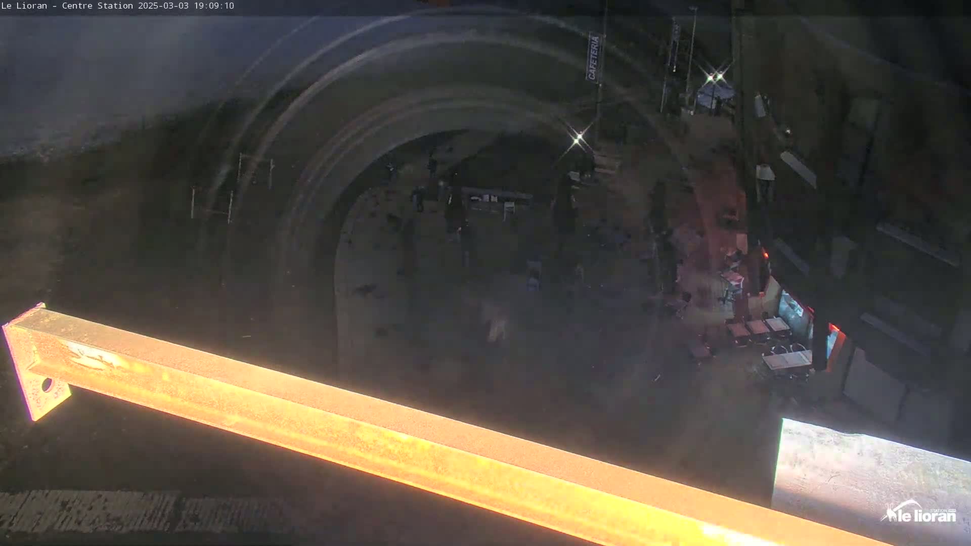 A high-angle, nighttime view shows a crowd of people gathered in a dimly lit area beneath a curved structure, near a cafeteria with outdoor seating.