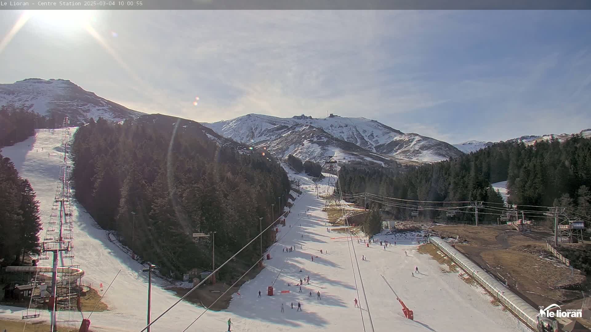 A sunny day at a snow-covered ski resort shows several ski slopes with skiers and snowboarders, ski lifts, and surrounding evergreen trees and snow-capped mountains.
