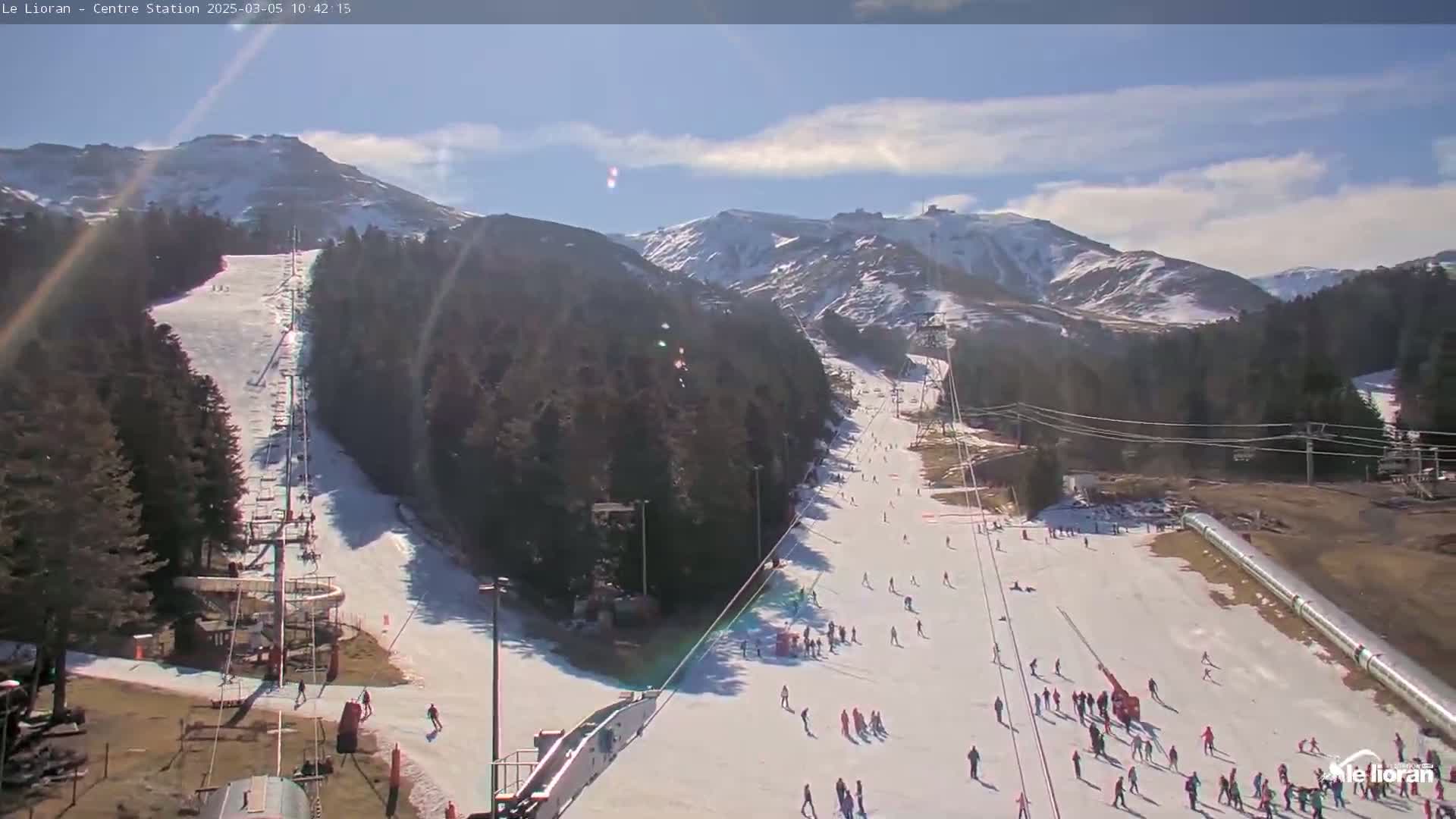A sunny day at a snow-covered ski resort shows several skiers and snowboarders on a slope, with mountains in the background and ski lifts visible.
