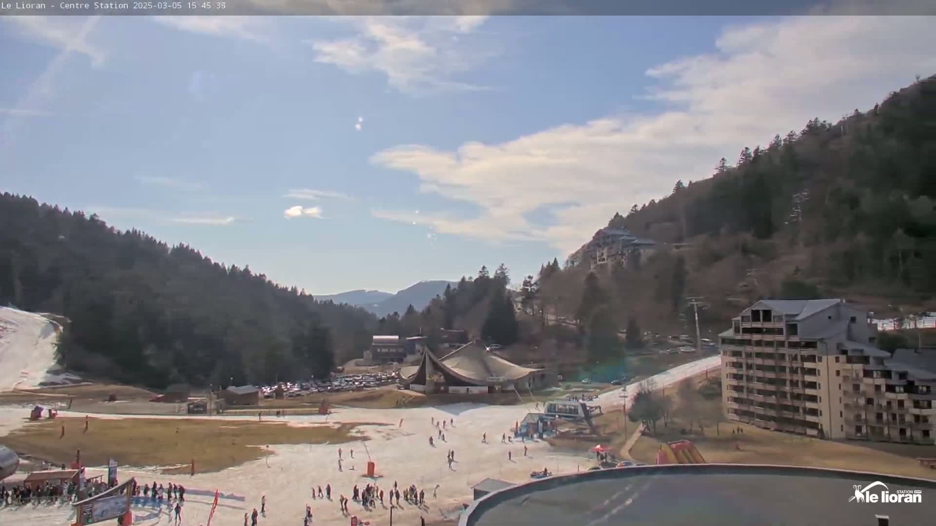 A partly sunny day reveals a ski resort nestled in a valley between snow-dusted mountains, with skiers scattered across the slopes and buildings visible nearby.