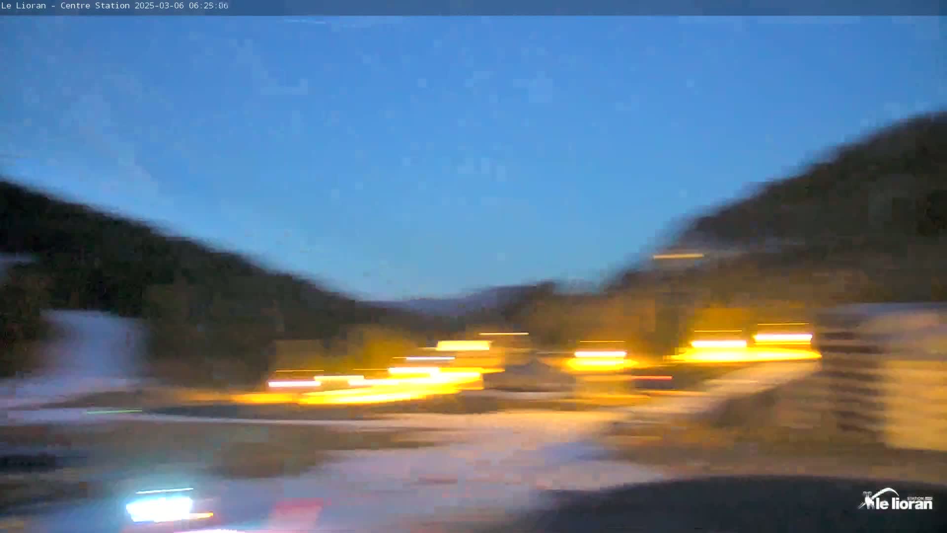 Under a twilight sky, blurred lights from moving vehicles illuminate a snow-covered road nestled between dark hills.