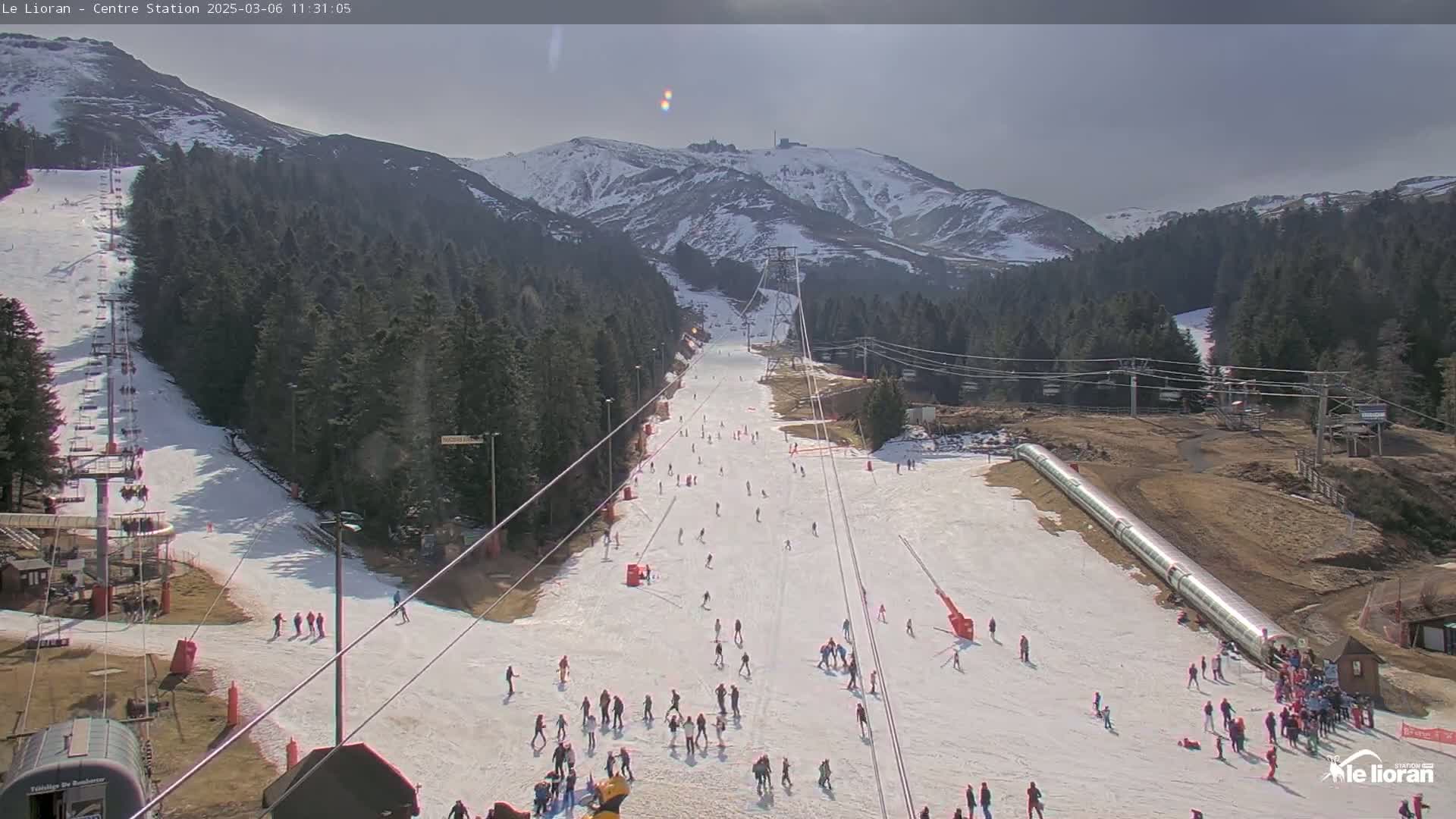 A ski slope with many skiers and snowboarders on a sunny day, surrounded by snow-capped mountains and evergreen trees.