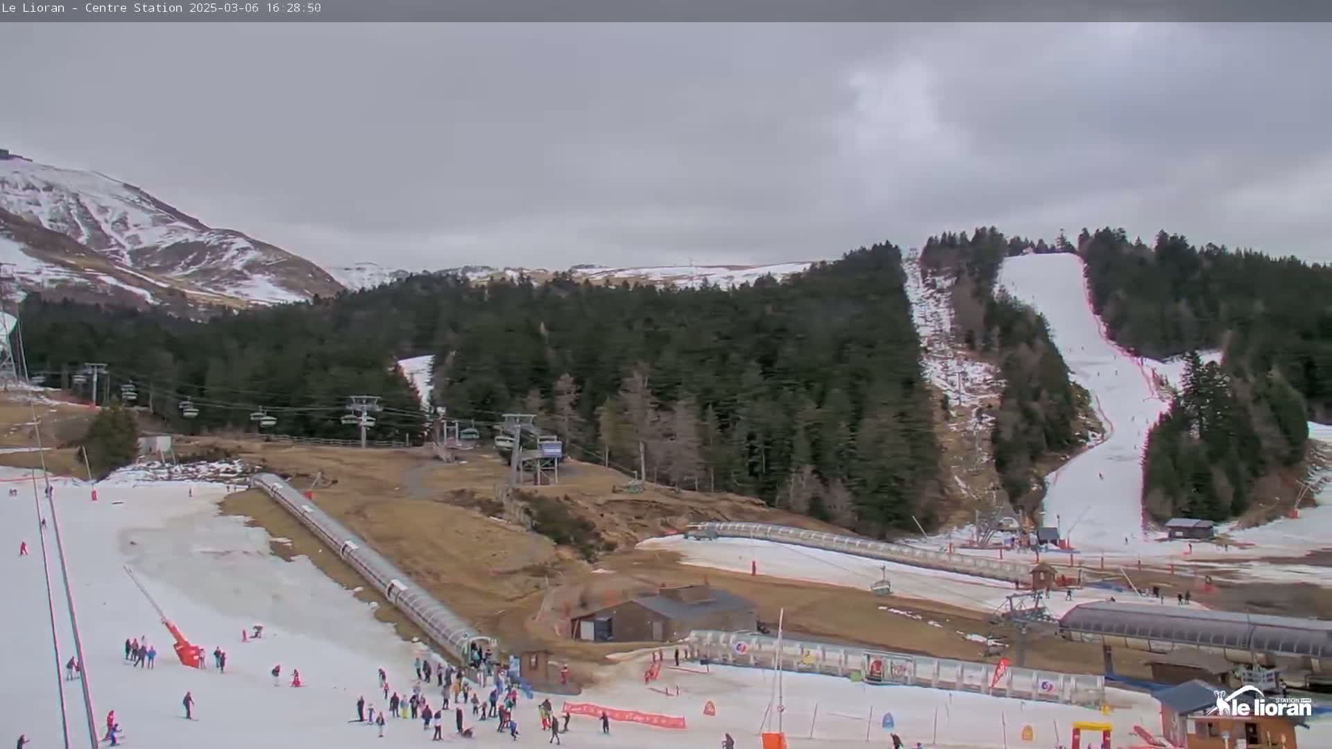 A ski resort with snow-covered slopes and hills under an overcast sky, showing several ski lifts, skiers, and buildings.