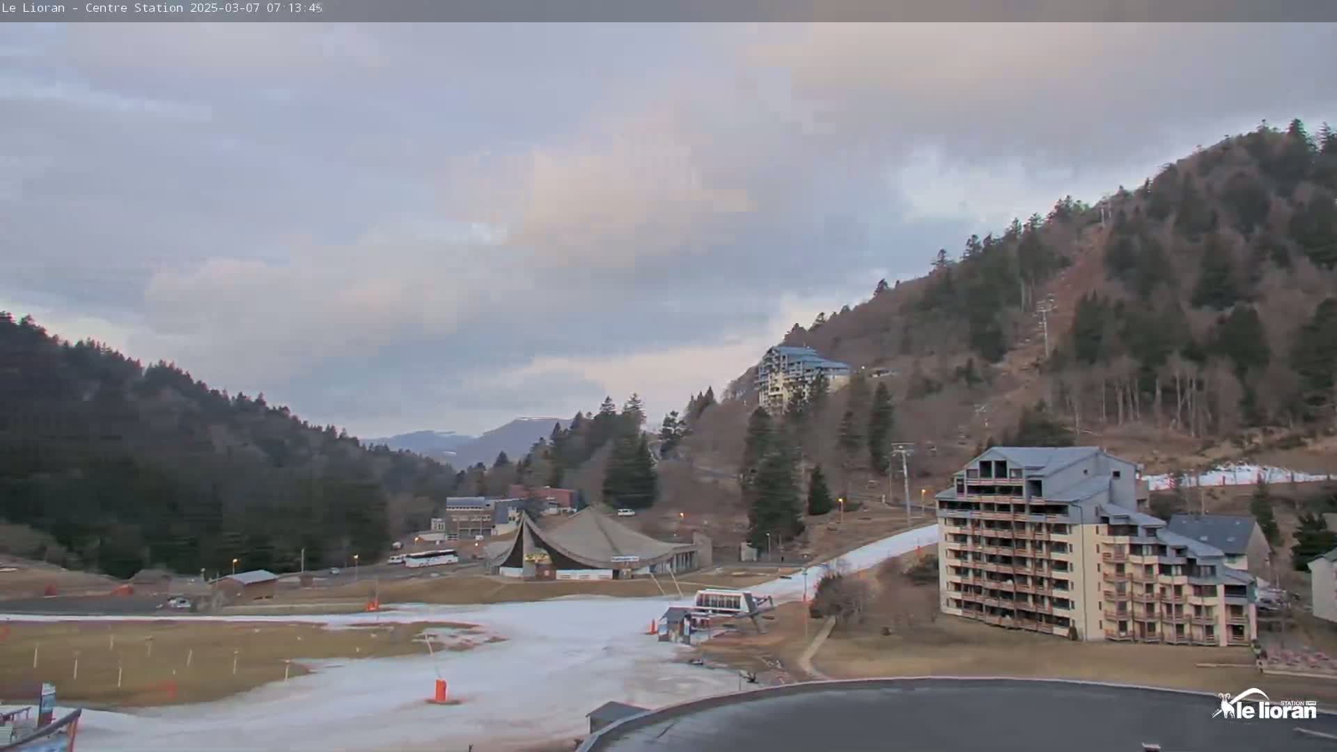 A mostly cloudy day reveals a ski resort nestled in a mountain valley, with snow visible on the lower slopes and buildings including hotels and a large structure.