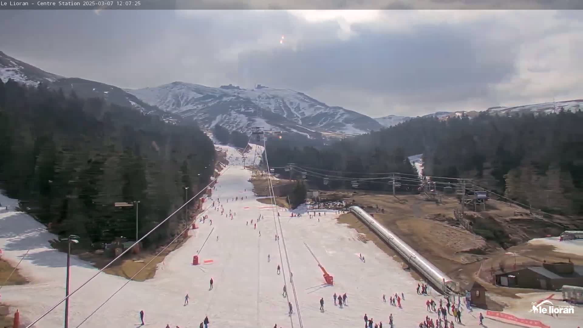 A ski slope with scattered skiers is seen beneath a mostly cloudy sky, with snow-covered mountains in the background.