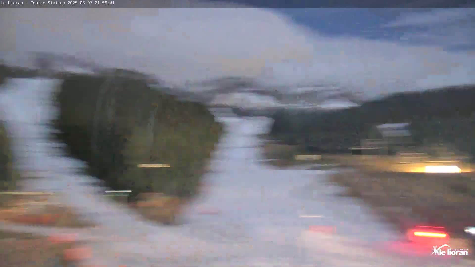 A blurry image shows a nighttime, snowy scene of a road with vehicles and dark hills in the background.