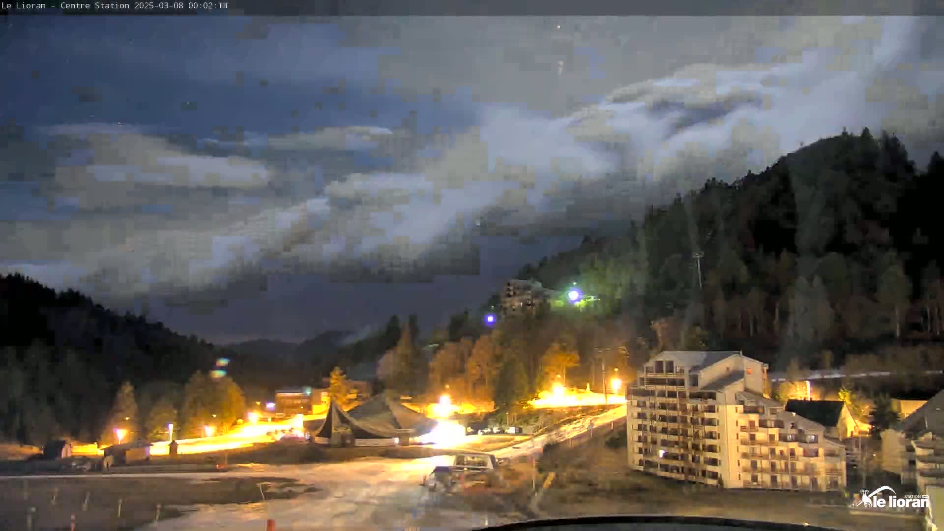 A nighttime view of a snow-covered mountain village under a cloudy sky, with lights illuminating buildings and roads.