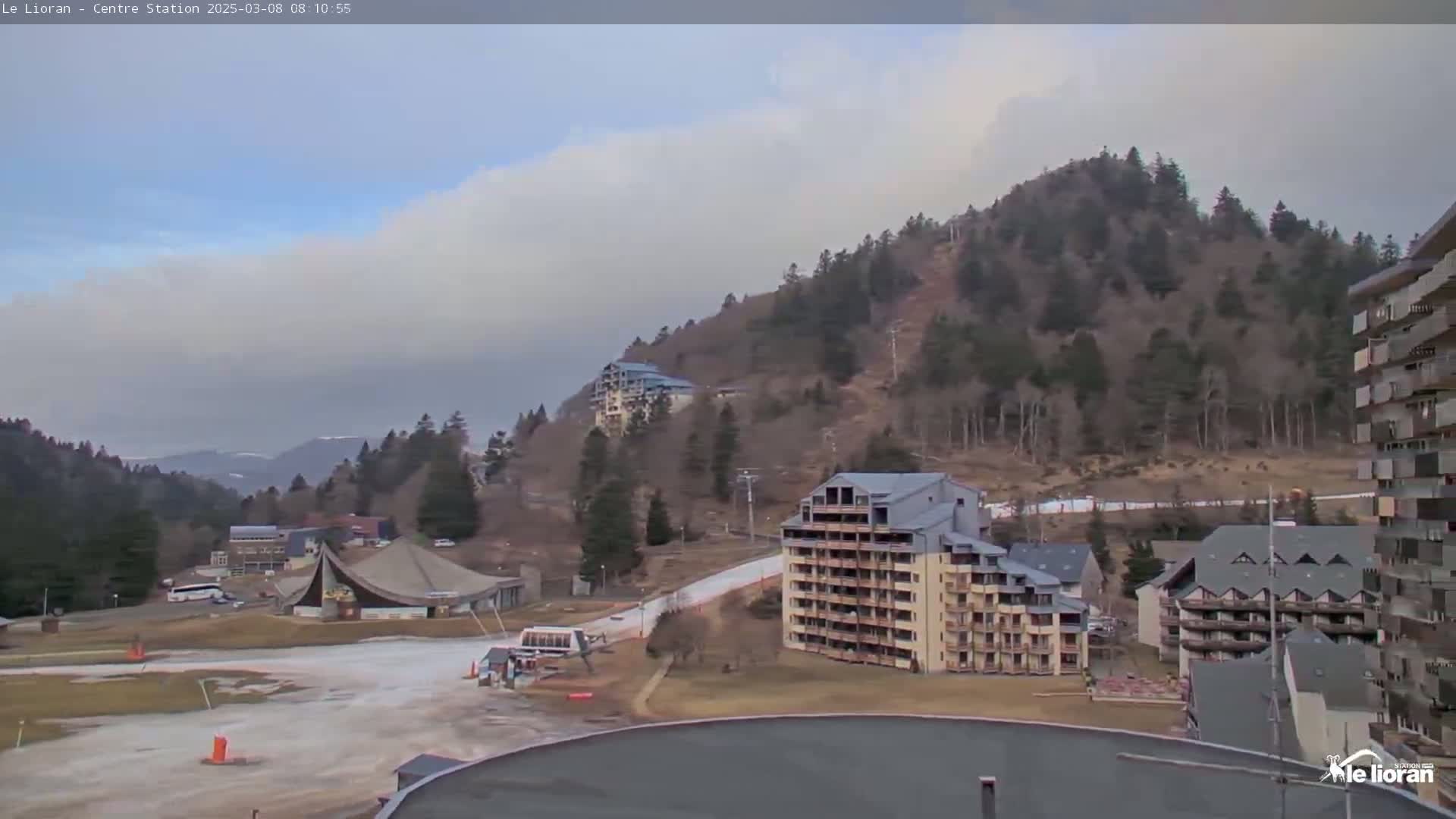 A cloudy day reveals a ski resort nestled among snow-dusted hills and buildings, with a flat area in the foreground.